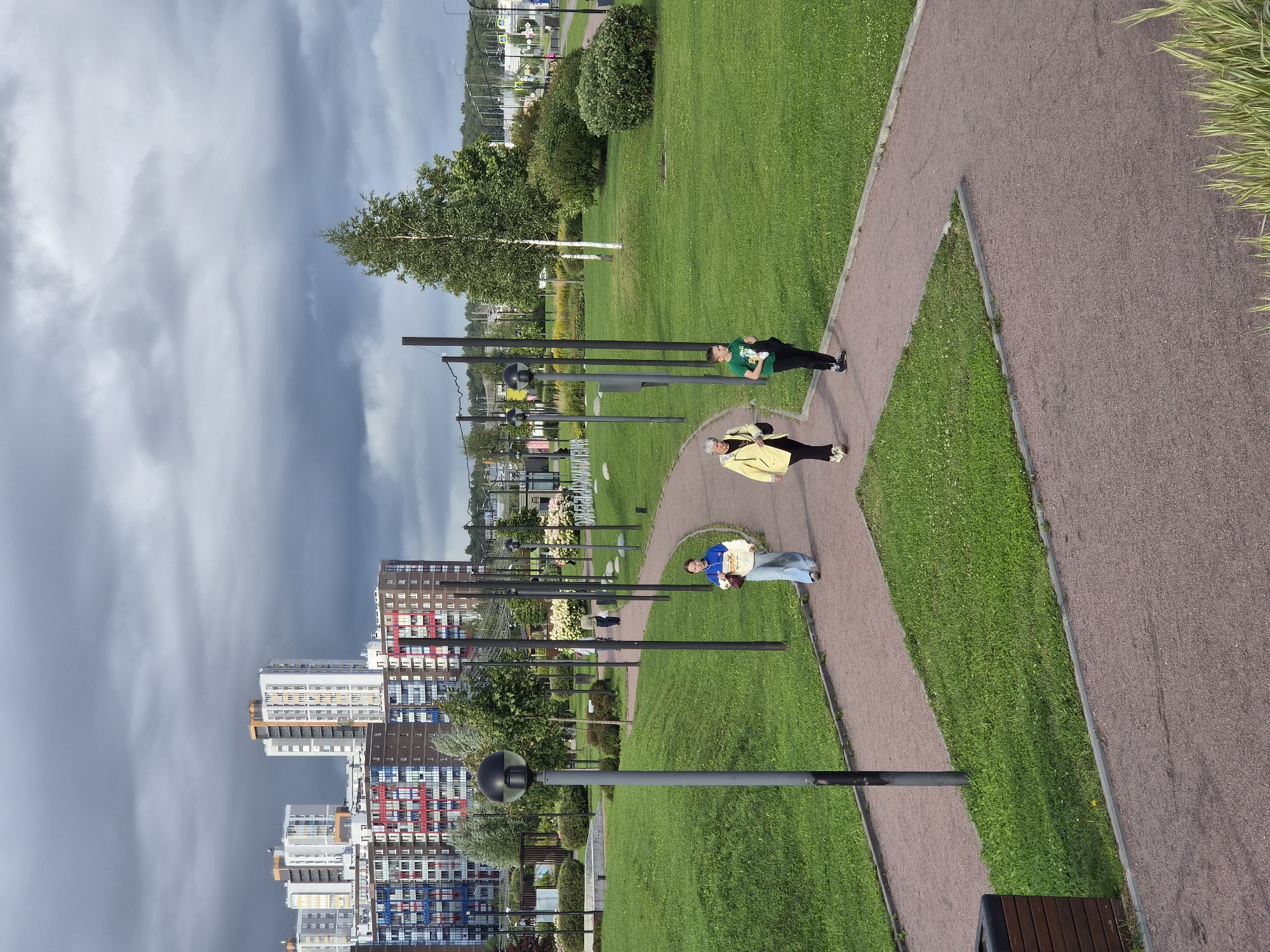 In Kudrovo, Russia, a paved pathway curves through a manicured green park. The pathway is a light reddish-brown color, contrasting with the vibrant green grass that surrounds it. Several tall, thin lampposts are strategically placed along the path, their dark poles and spherical lights creating a modern aesthetic. A few trees are scattered throughout the park, with a notable birch tree standing tall in the mid-ground. In the background, a cluster of multi-story apartment buildings dominates the skyline, their facades a mix of white, grey, and colorful accents, suggesting a residential area. The sky above is overcast with dark, dramatic clouds, hinting at potential inclement weather. Three individuals are walking along the pathway. In the foreground, a young person in a blue hoodie and light blue jeans walks away from the camera. Further along the path, an older woman in a bright yellow jacket and dark pants walks towards the camera, accompanied by a young person in a green t-shirt and dark pants. The overall scene depicts a well-maintained urban park with modern architecture in the background and a somber sky.