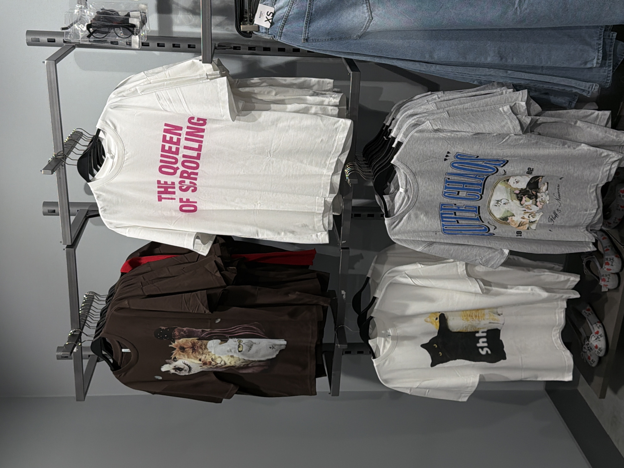 The image shows a retail display of graphic t-shirts and denim jeans within an indoor store. On the upper rack, there are several t-shirts hanging on hangers. The leftmost t-shirt is brown with a graphic of multiple cats. Next to it is a white t-shirt with pink text that reads "THE QUEEN OF SCROLLING." Behind these, partially visible, are more t-shirts and a red garment. On the lower rack, a white t-shirt with a graphic of two cats and the word "shh" is displayed prominently. To its right, there are several grey t-shirts with graphics featuring dogs and text. To the right of the lower rack, a pair of light-wash denim jeans are hanging, with an "XS" size tag visible. Two pairs of flat shoes with red and white patterns are placed on a shelf beneath the lower rack. The background is a plain grey wall. There are no people visible in the image. The scene depicts a retail environment, likely a clothing store, with merchandise neatly arranged on display racks. The presence of generic store fixtures and the organized presentation of clothing suggest a commercial setting. The overall impression is one of a well-stocked and clean retail space.