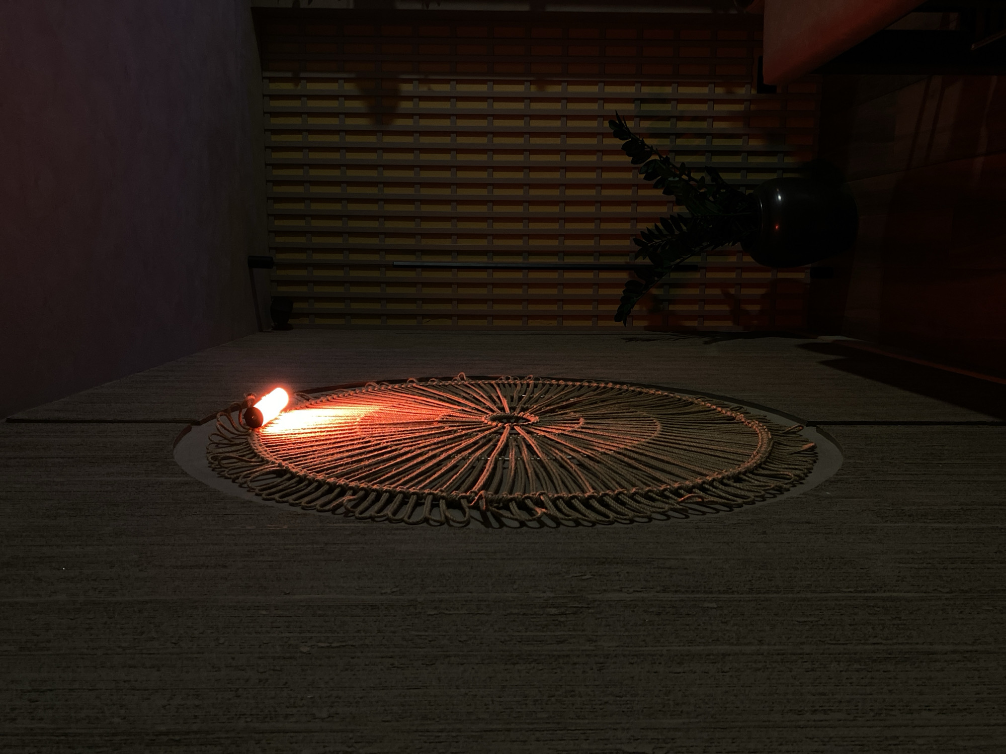 A low-angle, dimly lit photograph captures a round, decorative mat illuminated by a warm, orange light source, potentially a bulb or fire. The mat features intricate concentric circles formed by woven material, possibly rope or straw. It is set into a surface that appears to be a dark-colored platform or floor.
Behind the mat, a vertical wall with horizontal wooden slats is visible. To the right, a potted plant with dark green foliage is positioned near a dark, perpendicular structure. The location is Izhevsk, Russia.