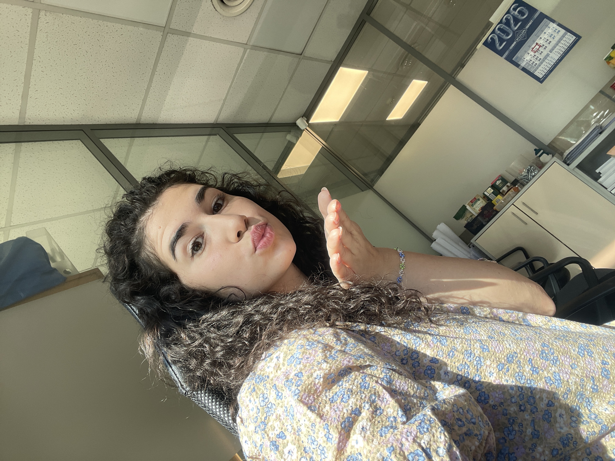 A young woman with long, curly, dark hair is taking a selfie in an office environment. She is wearing a light-colored floral print top with blue and pink flowers. Her left hand is extended towards the camera, and her lips are pursed in a kissing gesture. She is wearing a beaded bracelet on her left wrist with green, blue, and clear beads. In the background, there are glass partitions, a suspended ceiling with fluorescent lighting, and office furniture. A calendar for the year 2026 is visible on the wall. The scene suggests an indoor office setting, possibly in a commercial building. The presence of a calendar with "2026" is a clear indicator of the time frame. The overall lighting indicates it is daytime, with sunlight streaming in from the left, casting shadows. The location is an office in Sofia, Bulgaria.