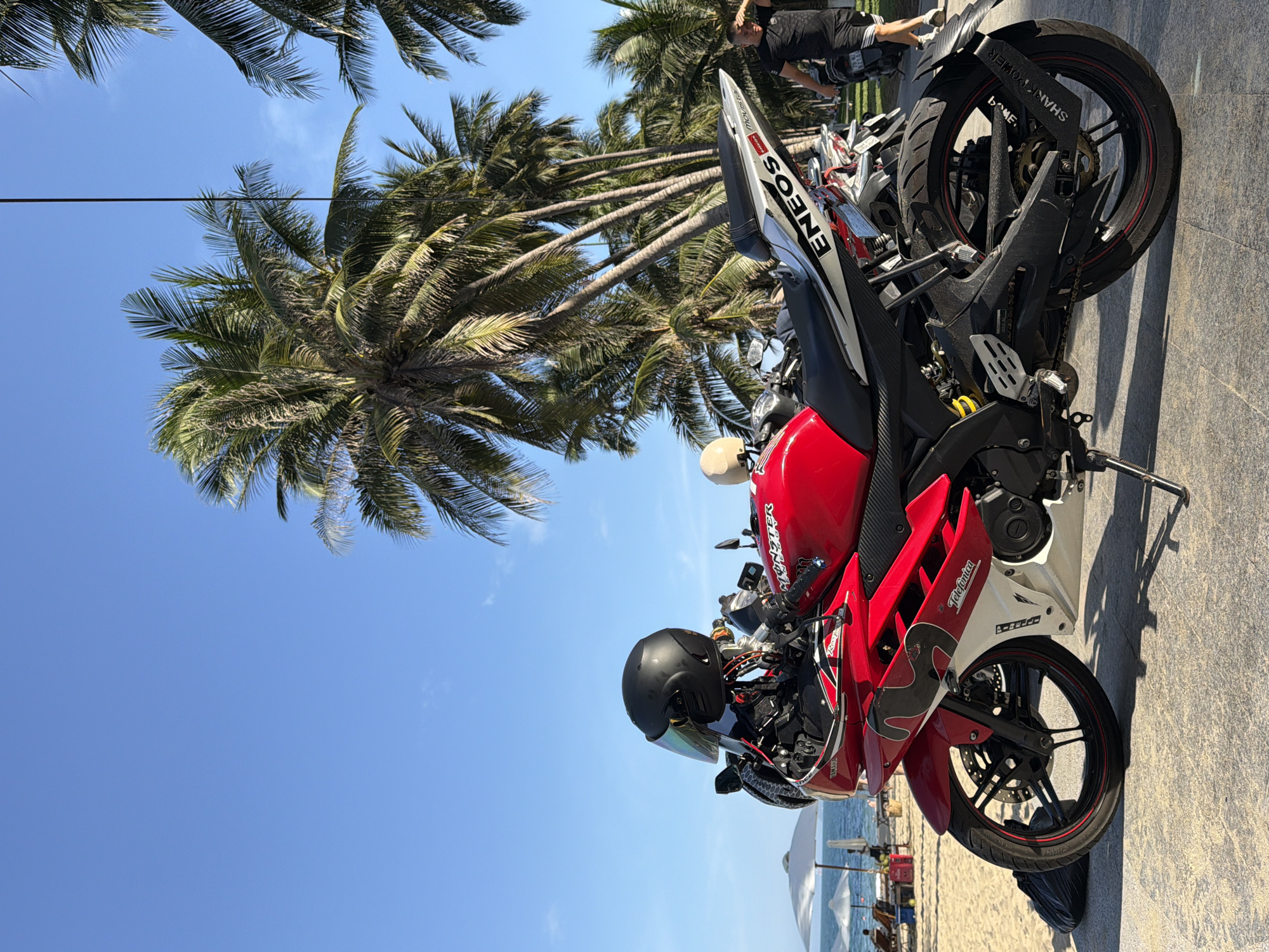 A red and white motorcycle is parked on a paved walkway next to a sandy beach in Nha Trang, Vietnam. The motorcycle has a black helmet with a tinted visor resting on its handlebars. In the background, palm trees line the beach, and the ocean is visible under a clear blue sky. Beach chairs and umbrellas are set up on the sand, with a few people in the distance. A man in a black t-shirt and shorts is walking away from the motorcycle towards the right side of the image.