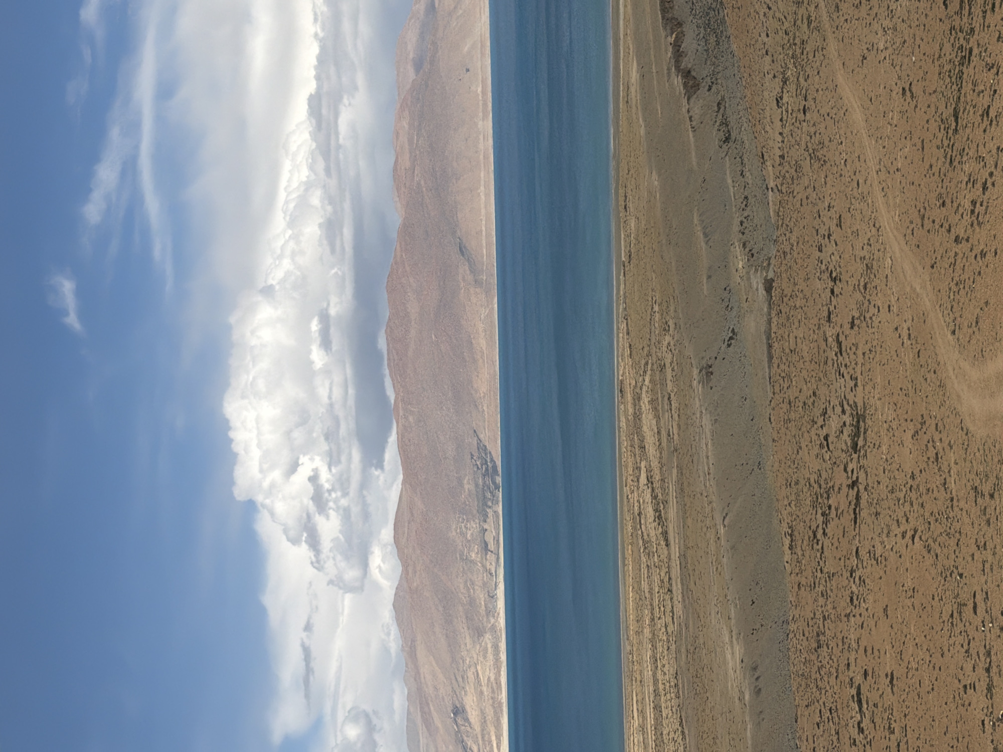 The image presents a landscape featuring a stark contrast between elements. On the left, a bright blue sky transitions into a bank of voluminous white clouds. Adjacent to this is a mountain range, with a tan and rocky texture. A body of water, dark blue, separates the mountains from a flat, barren expanse of brown, sandy terrain. The image's orientation is tilted. The exact location within Gyirong County is not identifiable from this image. There are no people visible in the image.