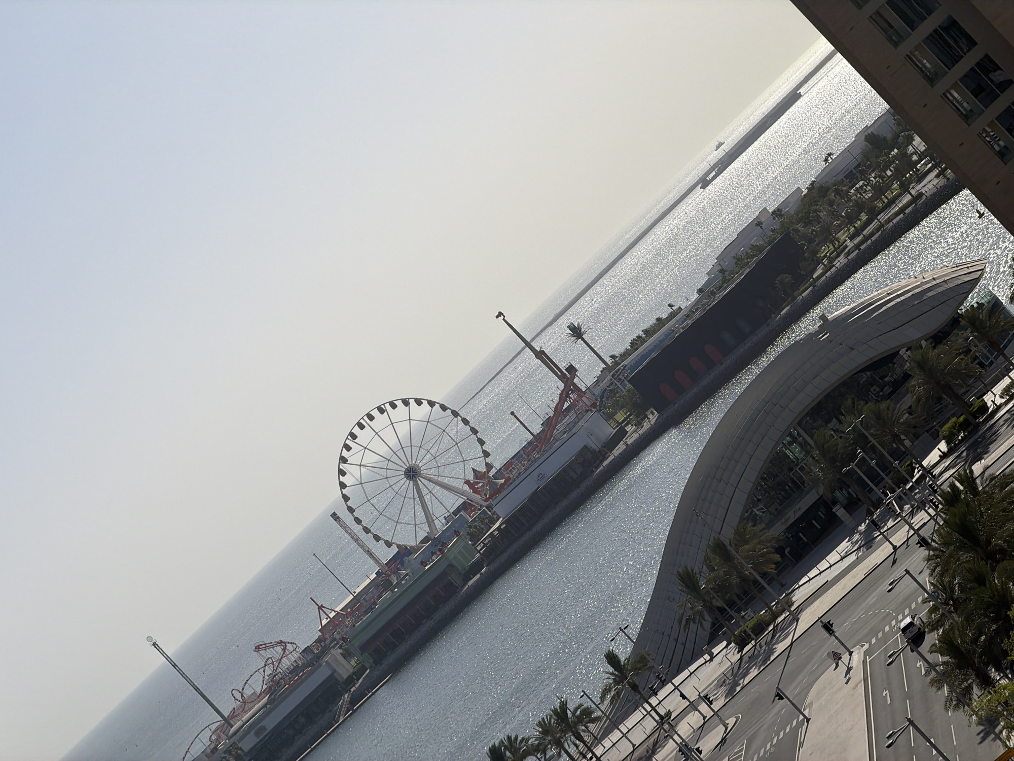 The image shows an aerial, tilted view of a waterfront area in a city. Dominating the scene are several large amusement park rides, including a prominent Ferris wheel and a roller coaster. The rides are situated along a pier or boardwalk that extends into a body of sparkling blue water. In the background, the water stretches to the horizon, with a pale, hazy sky above.
To the left of the waterfront, a modern building with a curved, glass facade is visible, surrounded by palm trees. A street with traffic lights and a white car is also present, indicating an urban environment. The overall scene suggests a recreational area within a city, likely a tourist destination or entertainment district. The lighting suggests it is daytime, with the sun reflecting off the water.