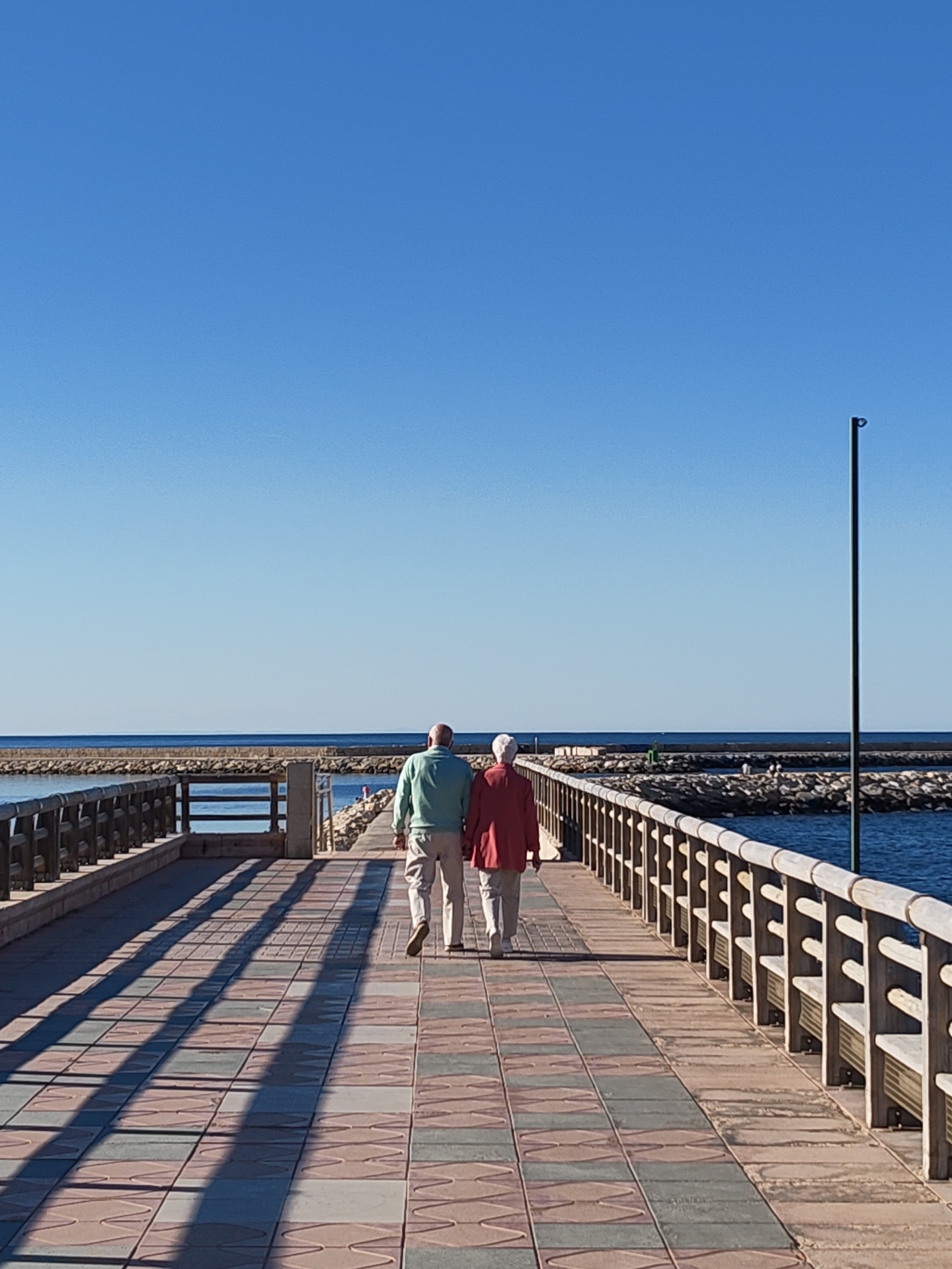 The image depicts an elderly couple walking hand-in-hand away from the viewer along a tiled pier under a vast, clear blue sky. The setting is a sunny coastal area in Dénia, Spain.
The pier features a pathway paved with patterned tiles in shades of red, green, and grey. Wooden railings with benches integrated into their design line both sides of the pier, casting long, pronounced shadows across the pathway, indicating either morning or late afternoon.
To the left of the couple, the pier runs alongside a rocky breakwater, beyond which calm, blue water is visible. To their right, the pier extends into the open sea, with a green navigational buoy or marker visible in the distance. A tall, thin black pole stands vertically on the right side of the pier.
The man, on the left, wears a light green long-sleeved shirt and light-colored trousers. The woman, on the right, is dressed in a red long-sleeved top and light-colored trousers, with white hair. They appear to be enjoying a peaceful, leisurely stroll together, their linked arms conveying a sense of companionship and affection. The overall atmosphere is serene and bright, characteristic of a pleasant day by the sea. No visible text is present in the image.