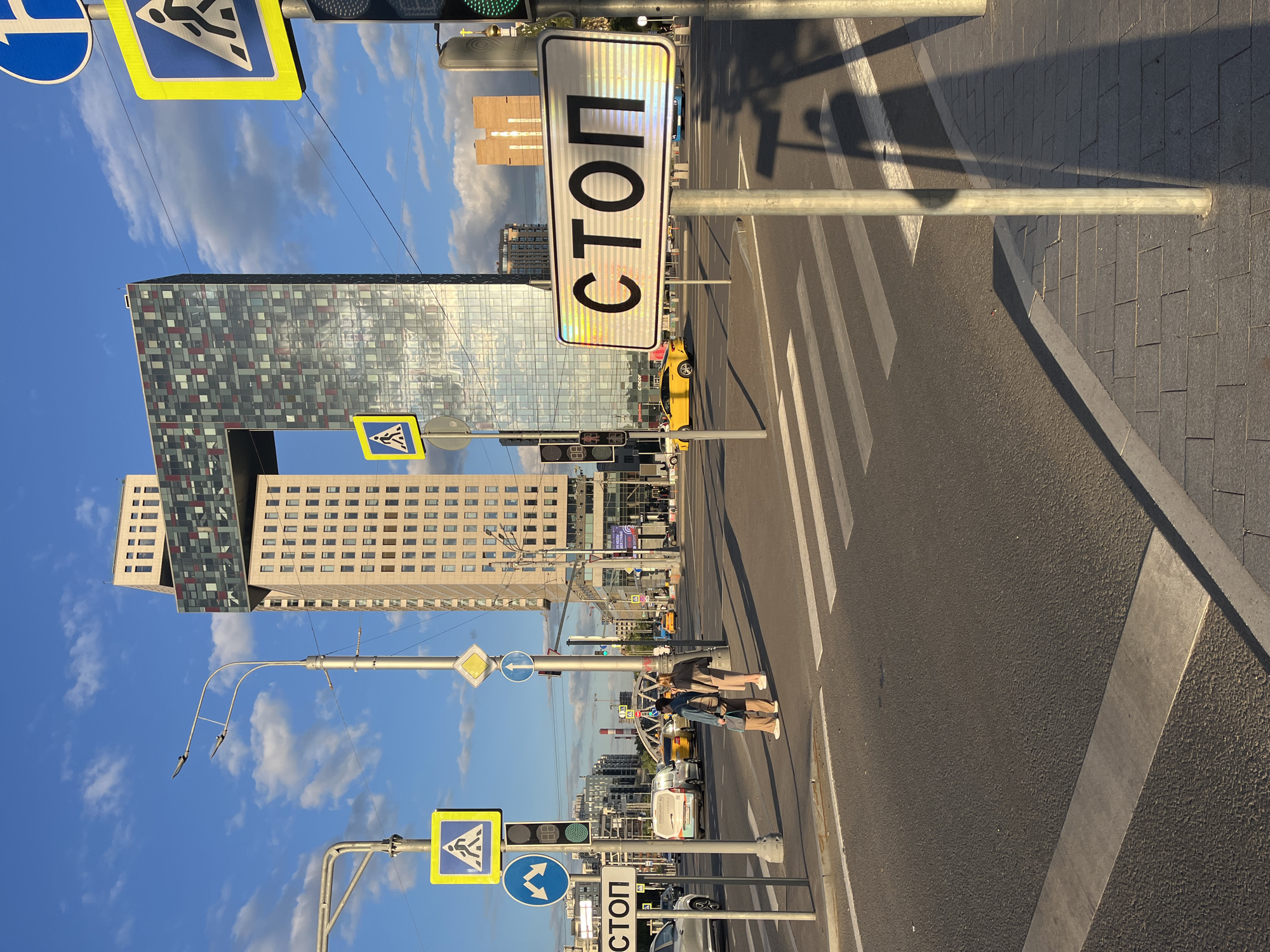 The image shows an urban street scene on a sunny day. Buildings of various architectural styles are visible in the background. In the foreground, there is a pedestrian crossing with two people walking across it. Traffic lights and road signs are present on either side of the road. The road surface is asphalt, and the pedestrian crossing is marked with white stripes. A yellow car is stopped nearby. A sign with the Russian word "СТОП" (STOP) is prominent. The location is likely in Russia, based on the sign's language.