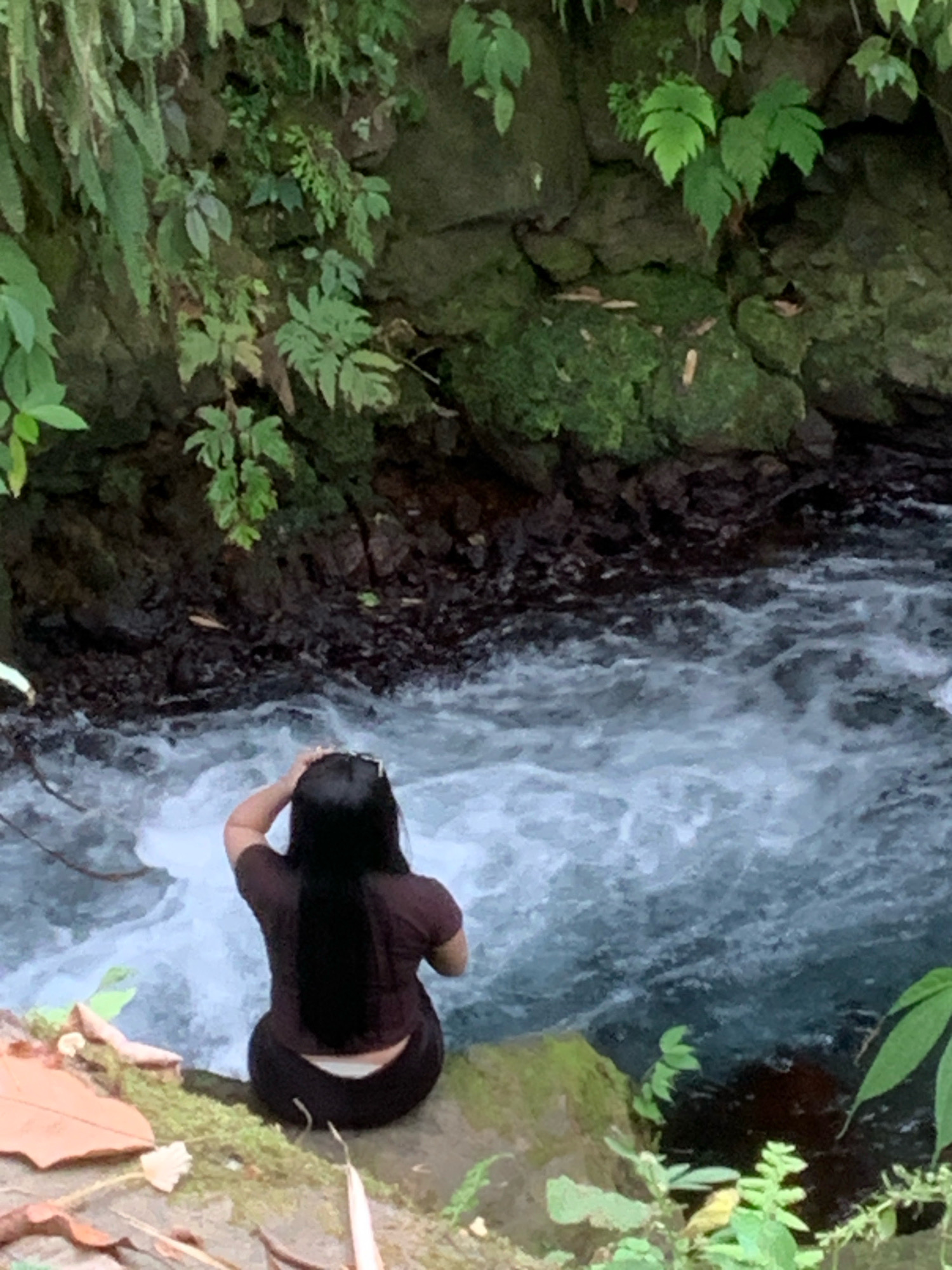 A person is seated on a moss-covered rock near a fast-flowing river in a lush, green environment. The person, viewed from behind, has long black hair and is wearing a dark brown t-shirt and dark pants, with white underwear visible at the waist. They are reaching up to touch their hair. The river has turbulent, white-capped water. The background consists of rocky cliffs covered in dense vegetation, including ferns and other leafy plants. The location is identified as Pronojiwo, Indonesia.