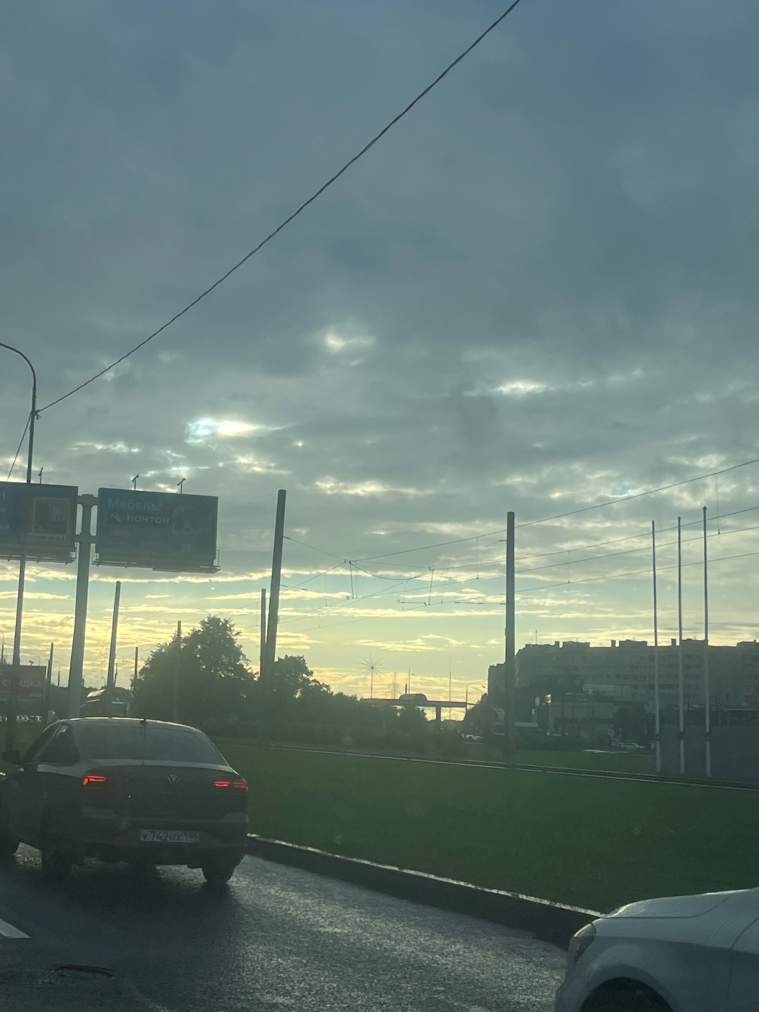 The image depicts a view from a vehicle, likely on a street in Saint Petersburg, Russia, during overcast weather with some sunlight breaking through the clouds. In the foreground, the rear of a dark sedan is visible, with its license plate reading "E742XX98". The road surface appears wet, suggesting recent rain. To the left, a tall street lamp is present. In the mid-ground, there are two large billboards. The left billboard is partially obscured, but the right billboard displays Russian text, including "МЕБЕЛЬ" (furniture) and "НоНТОН". Beyond the billboards, trees are visible, silhouetted against the bright sky. In the background, a cityscape with several multi-story buildings and what appears to be a bridge or overpass can be seen. Overhead, there are power lines and poles. The overall impression is a common urban scene with a focus on traffic and infrastructure under a dramatic sky.