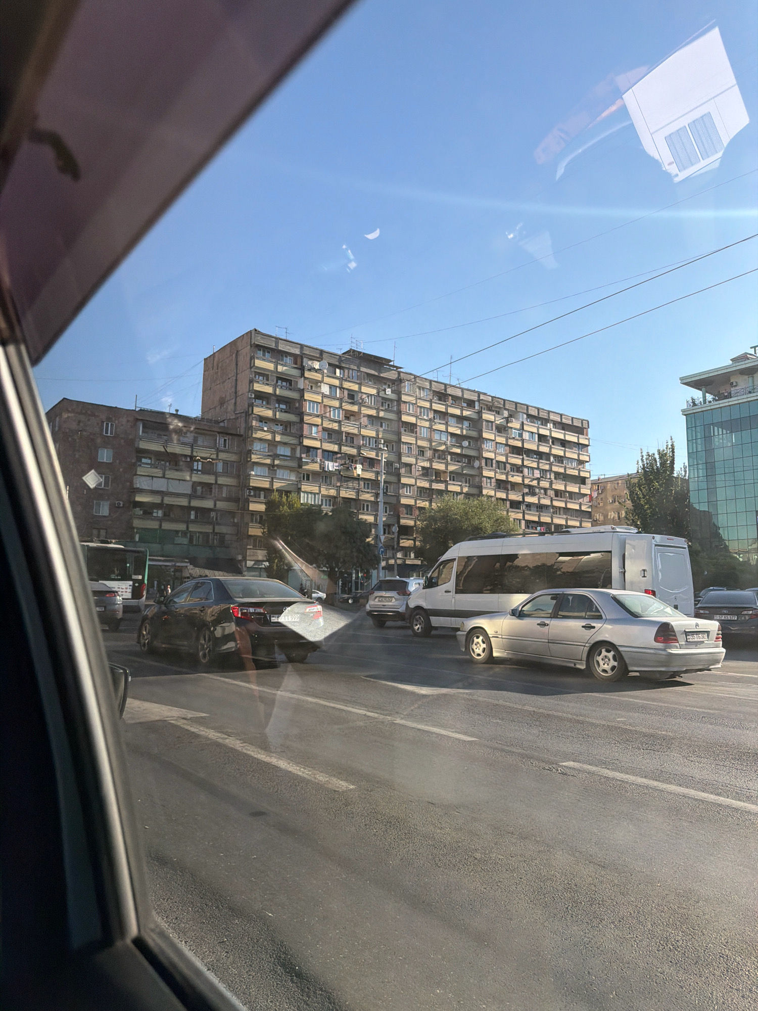 The image captures a street scene in Yerevan, Armenia, viewed from inside a vehicle. A large, Soviet-era apartment building with numerous balconies and windows dominates the background. In the foreground, traffic is visible, including a white van and several cars. A silver sedan is prominently featured in the middle ground. The sky is clear and blue, with a faint crescent moon visible. The overall impression is of a typical urban environment in Yerevan, characterized by its distinct architecture and active street life.