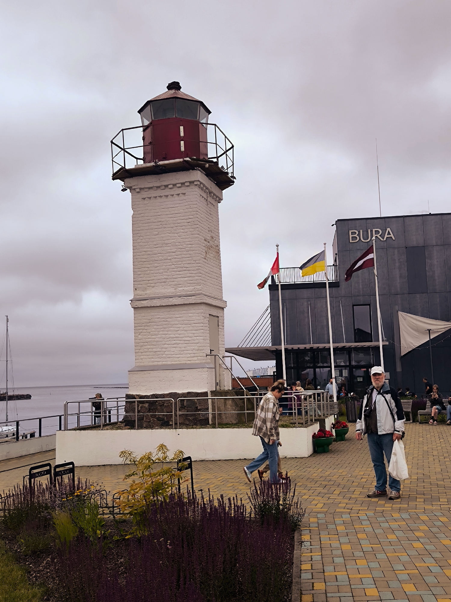 The image captures a waterfront scene in Salacgrīva, Latvia, with a prominent lighthouse and a modern building. The lighthouse, white with a red top, stands to the left, its brickwork weathered. To the right, the building, identified by the "BURA" signage, features a dark, textured facade. Three flags, including the Latvian flag, fly in front of the building. Several people are visible: a man in a white cap and jacket stands in the foreground holding a white bag, while a woman in a plaid shirt walks past him. Other individuals are scattered in the background, near the building entrance and along the waterfront. The ground is paved with a pattern of yellow, grey, and blue tiles. Lush purple and green vegetation lines the foreground. The sky is overcast, and the calm water of the sea extends to the horizon, with a sailboat visible in the distance.