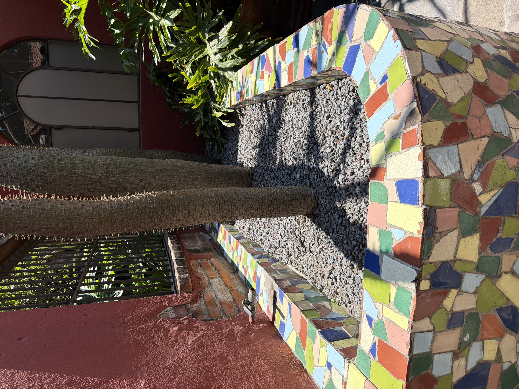 A photograph taken in Puebla, Mexico, showcases a garden element featuring a curved, mosaic-tiled wall or structure. The tiles are various colors, including blue, red, green, and yellow. Inside the mosaic structure, the space is filled with grey gravel. A cactus with a thick, spiky trunk extends across the structure. Beyond the cactus and gravel, the backdrop includes a building with a window and green foliage. Another section of the mosaic structure connects to a red-painted wall and has a metal spigot.