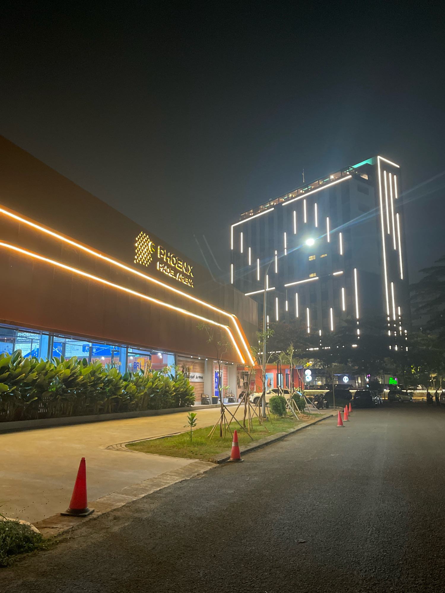 The image shows a nighttime view of a modern building complex in South Tangerang, Indonesia. The main building on the left has a dark brown facade illuminated by horizontal orange LED strips. A sign with "PHOENIX PADEL ARENA" in yellow letters is visible on this building. In front of it, a row of lush green plants lines the sidewalk.
To the right, a tall, multi-story building dominates the skyline, its facade adorned with numerous vertical white LED lights. The sky above is dark and clear. In the foreground, a dark asphalt road curves to the right. Several orange traffic cones are placed along the edge of the road and sidewalk, indicating some form of demarcation or construction. Parked cars and motorcycles are visible in the background, suggesting a public or commercial area. The overall impression is one of a contemporary urban environment at night, well-lit and seemingly active.