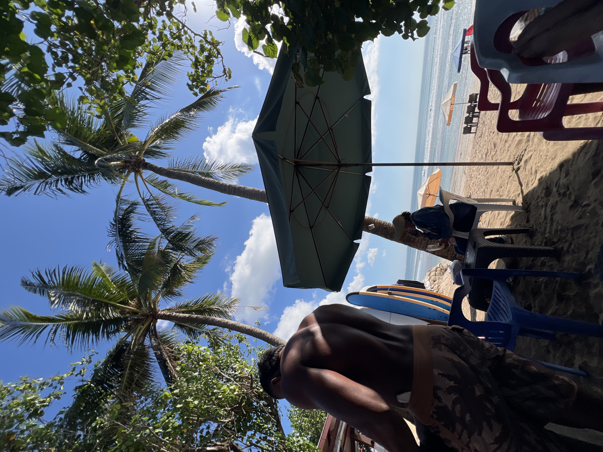 In Kuta, Indonesia, on a sun-drenched beach, a man with dark skin and muscular physique stands facing away from the camera, his back illuminated by the sun. He wears camouflage-patterned shorts. Beside him, leaning against a wall or structure, are two surfboards, one blue and yellow, the other with a design that is difficult to discern. The scene is set under the shade of palm trees and a large, dark green beach umbrella, which casts shadows on the sandy ground.
In the background, a beach stretches towards the ocean, with gentle waves lapping the shore under a bright blue sky dotted with white clouds. Several beach chairs and umbrellas are scattered across the sand, some occupied by people. To the left, another person, possibly a woman wearing a hat and a blue jacket, sits at a table. Further down the beach, a row of lounge chairs faces the ocean, with two peach-colored umbrellas providing shade. A red and blue umbrella is also visible near the water's edge. The overall atmosphere suggests a relaxed beach setting.