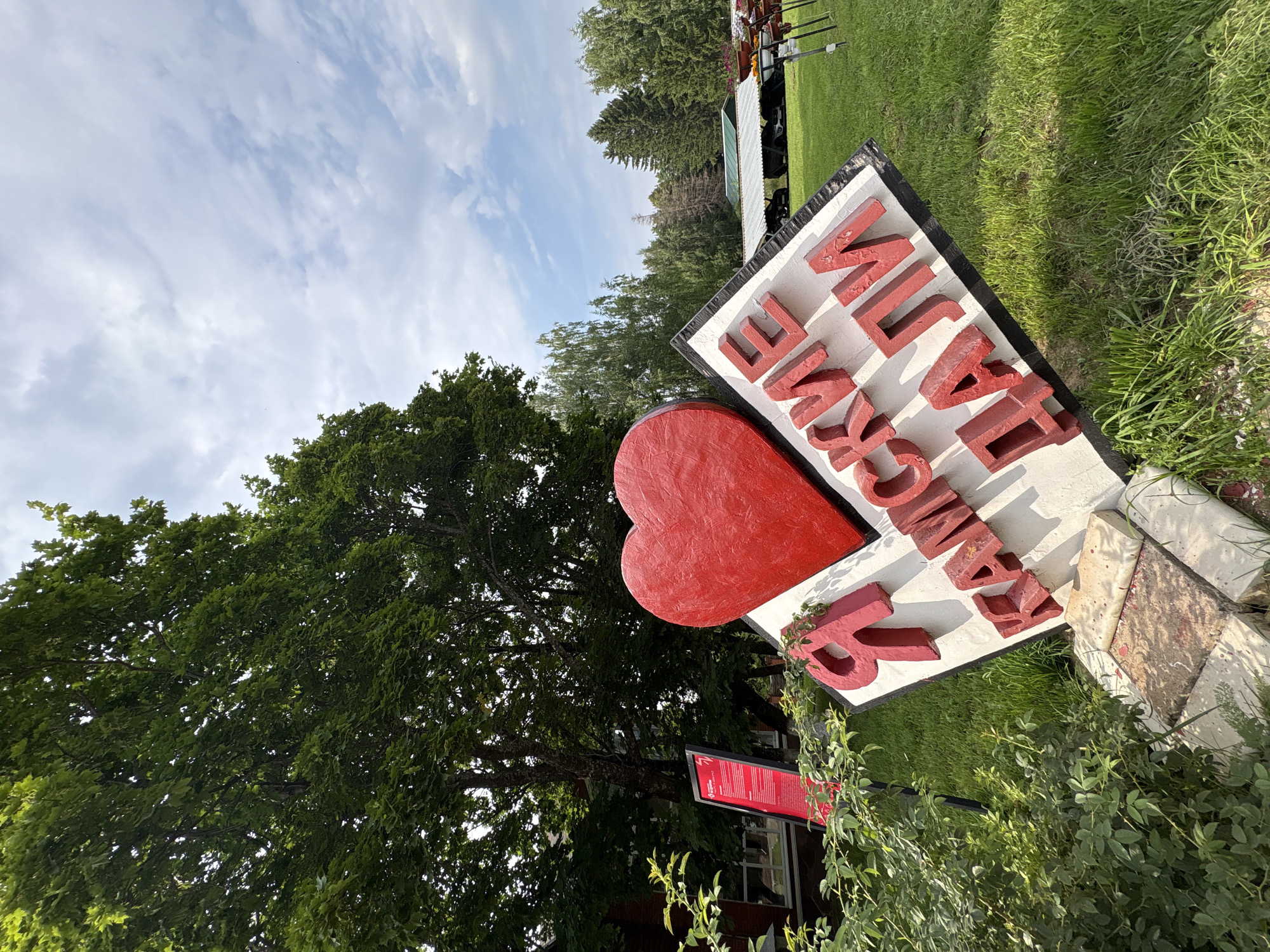 An eye-level, medium shot captures a large, red heart symbol positioned above a sign with red Russian lettering, set against a backdrop of a park-like environment. The sign, tilted at an angle, reads "Я КАМСКИЕ ДАЛИ," which translates to "I am Kama's expanses." The letters are three-dimensional and red, contrasting with the white background of the sign. The heart is solid red and appears to be made of a textured material. Surrounding the sign is a patch of green grass, and in the foreground, leafy green bushes are visible. To the left of the heart and sign, a red information board with white text is partially visible. In the background, tall green trees dominate the scene, with a bright, partly cloudy sky overhead. Further in the distance, a covered structure with cars parked underneath can be seen, and rows of potted flowers on stands are also present. The overall impression is of an outdoor monument or art installation in a scenic location. This scene is located in the city of Galevo, Russia.