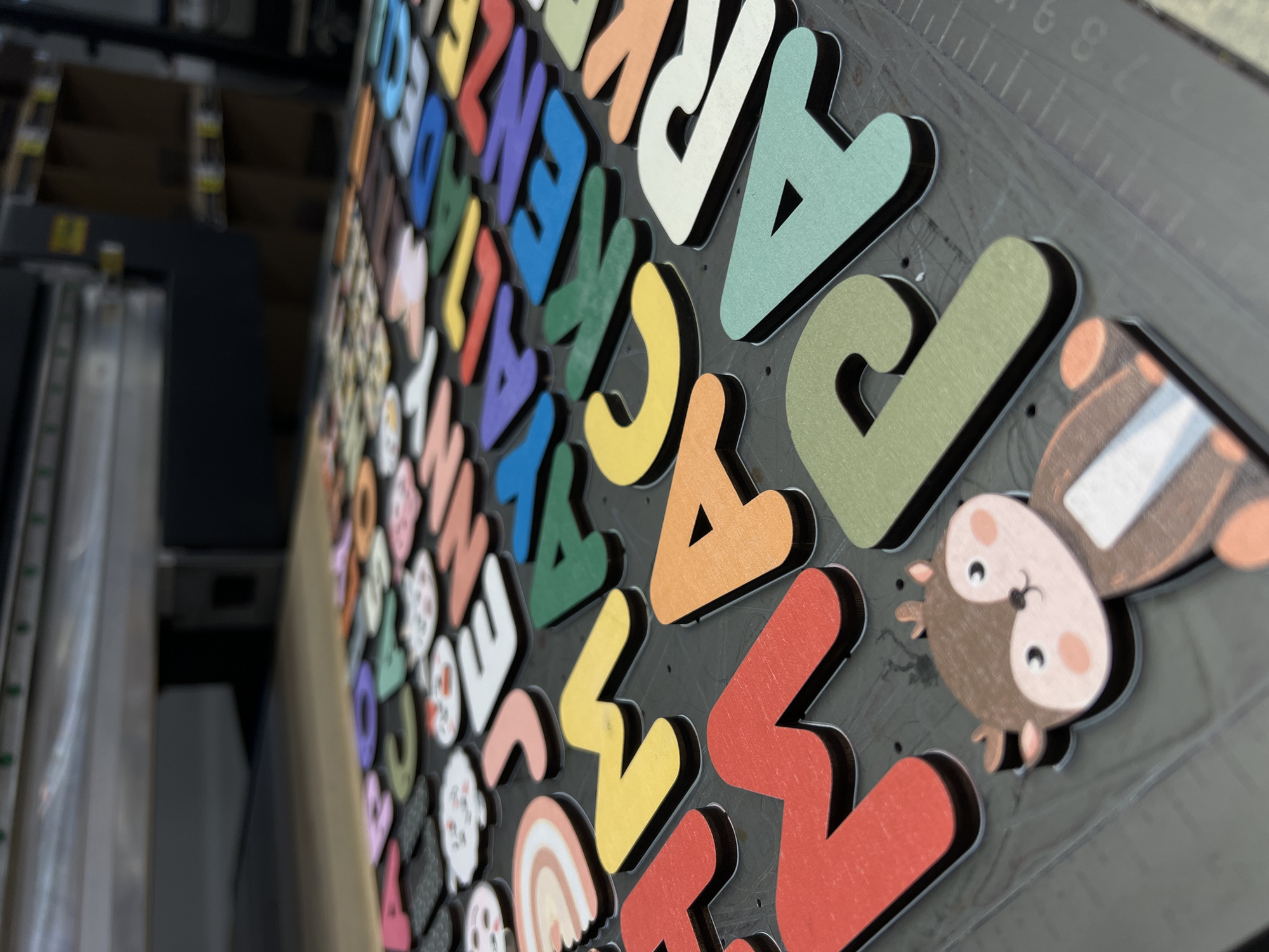 This image displays a close-up, angled view of a dark gray surface covered with colorful, three-dimensional wooden letters and several small, cartoonish animal figures. The letters are arranged in rows and columns, with each letter having a distinct color, such as blue, purple, green, yellow, orange, red, and white. The letters have a thick, black edge, giving them a layered appearance.
The animal figures appear to be a stylized owl and possibly other small creatures, also with bold outlines. One owl figure is prominent in the lower right quadrant of the image, with large eyes, pink cheeks, and brown and white coloring. Other smaller figures, possibly ghosts or snowmen with faces, are interspersed among the letters.
The surface beneath the letters has faint, gray markings resembling a ruler or grid pattern, with visible tick marks and numbers like "6", "7", "8", and "9", suggesting it might be a workbench or a prepared surface for crafting or manufacturing. In the blurred background, there are hints of machinery or equipment, possibly in a workshop or production environment. The lighting is bright and focused on the foreground elements, highlighting their textures and colors. There is no discernible text on the letters or figures themselves, other than the numerical markings on the background surface. The overall impression is of a collection of decorative elements, likely for educational or crafting purposes, in a production or assembly setting.