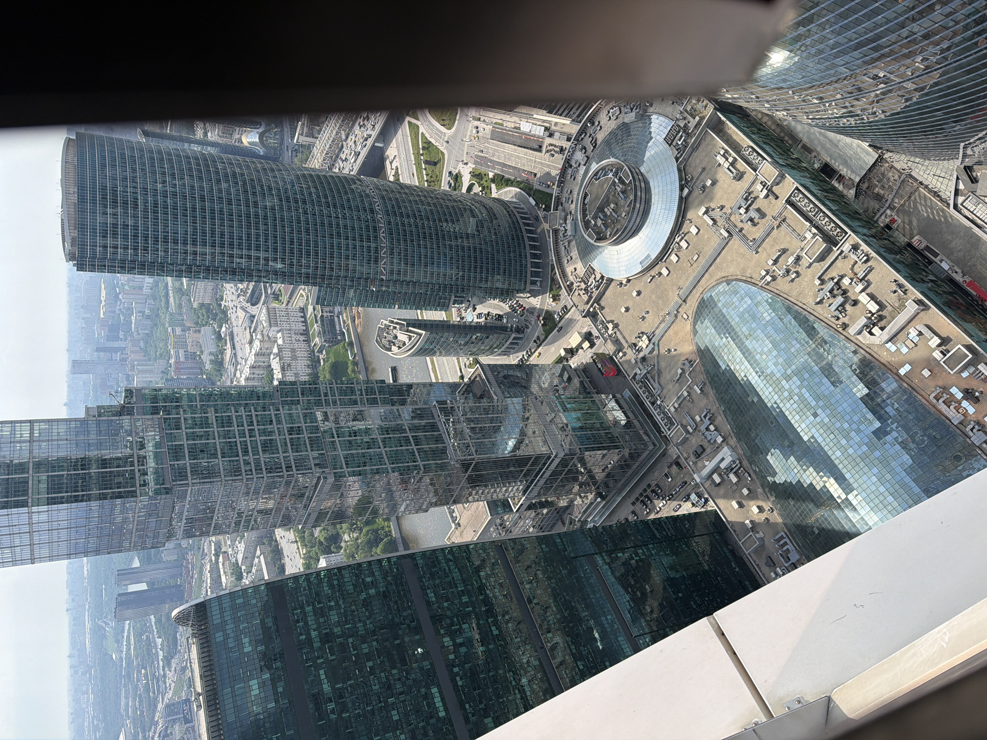 The image depicts a high-angle view of a city with modern architecture, possibly Khimki, Russia. Prominent are several skyscrapers with glass facades, indicating a dense urban area. The buildings' design suggests they are part of a modern business or residential complex. There's a circular structure, which could be a helipad or a decorative element, situated among the buildings. Roads and other urban infrastructure are visible, indicating a well-planned city layout. The sky is overcast or hazy, affecting the overall visibility. A section of a building's interior or balcony from where the photo was taken is visible in the foreground.