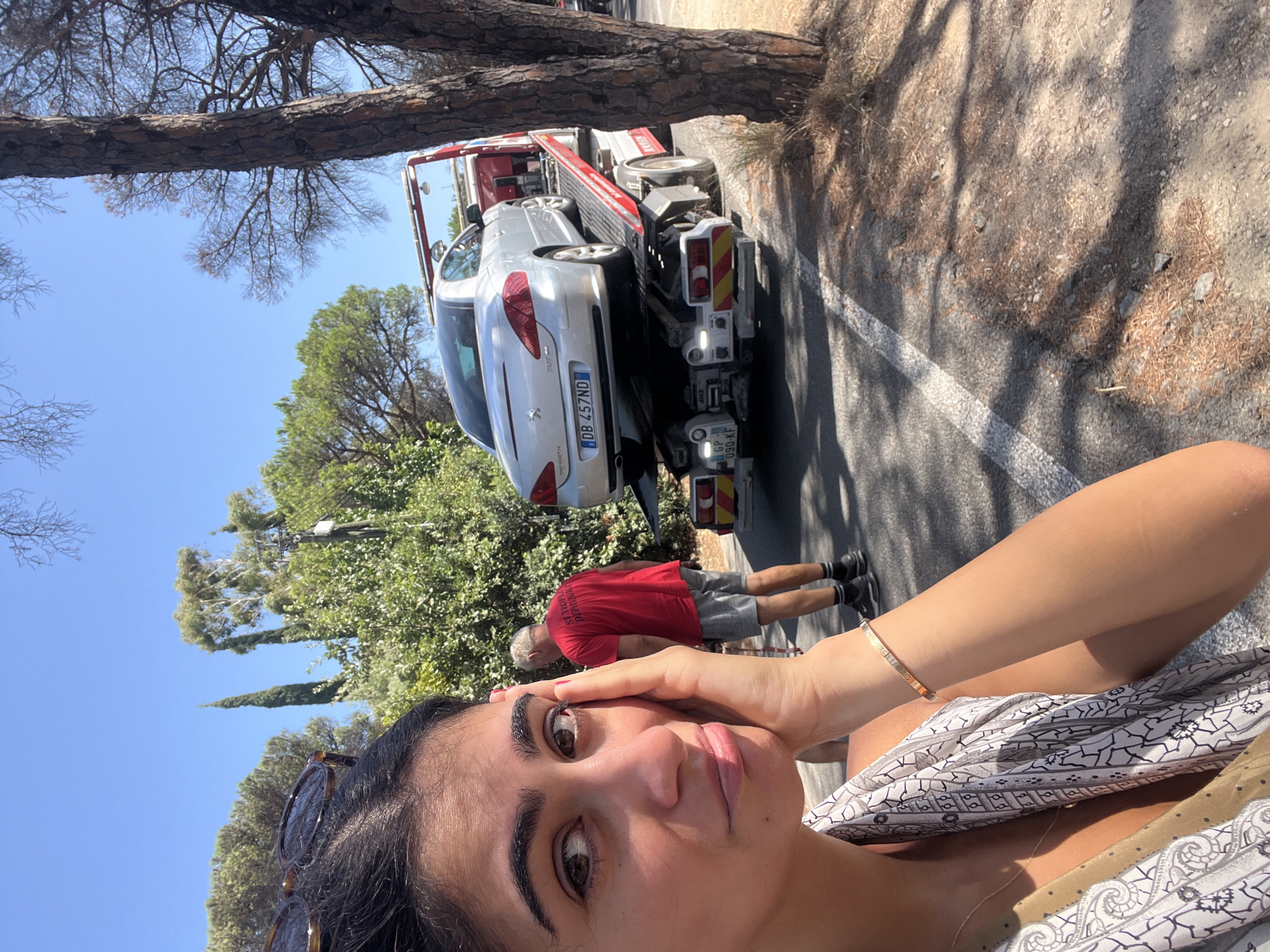 A young woman with dark hair and sunglasses perched on her head is looking at the camera in the foreground. She is resting her right hand on her cheek and wearing a patterned scarf. In the background, a silver Peugeot car is loaded onto a red tow truck. A man wearing a red t-shirt with the words "ST TROPEZ DEPAN..." printed on it and grey shorts is standing behind the tow truck. The scene takes place on a sunny day on a road bordered by trees and dry vegetation, likely in Ramatuelle, France, as indicated by the text on the man's shirt. The car's license plate is DB 457ND. The tow truck also has a license plate that appears to be GP 090-KF.