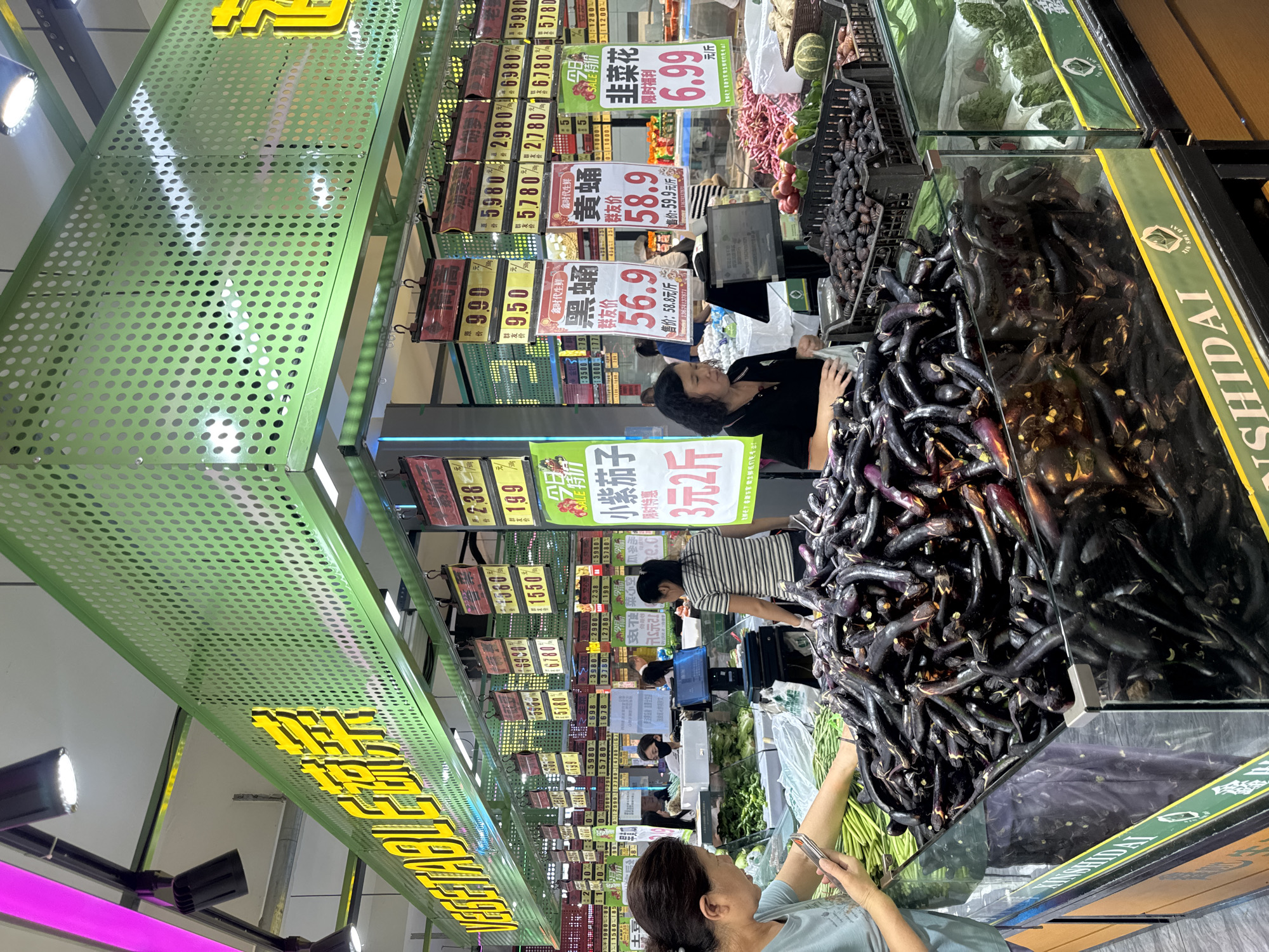 The image depicts a brightly lit indoor market scene, likely a supermarket produce section, in Shenyang City, China. A prominent green metal structure with circular perforations spans across the top of the frame, featuring a large yellow sign with Chinese characters, partially obscured by the text "VEGETABLE DU", suggesting it's a vegetable display area. Below this, various price tags are displayed, with some featuring Chinese text and numbers, such as "990", "950", "238", "199", "5980", "2980", "5780", "2780", and "6.99".
In the foreground, a large display of dark purple eggplants is piled high in a glass-encased bin. To the left, a woman in a light blue t-shirt is holding a smartphone and appears to be looking at it while standing next to a bundle of green beans. To her right, another woman in a striped t-shirt is interacting with the eggplant display. Further back, several other individuals are visible, some attending to the produce and others browsing. In the mid-ground, black plastic crates hold smaller, dark items, possibly insects or seeds, and other produce like red onions and gourds are visible in the background displays. The overall environment suggests a busy retail setting focused on fresh produce.
