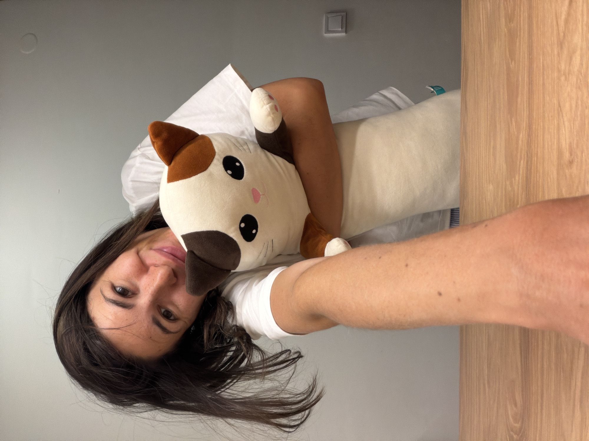A woman with dark hair is lying in bed and holding a plush cat toy. She is wearing a white t-shirt and is smiling slightly. The cat toy is brown and white with large black eyes. The background is a gray wall and a wooden surface. There is a light switch on the wall. There is no information about the location.