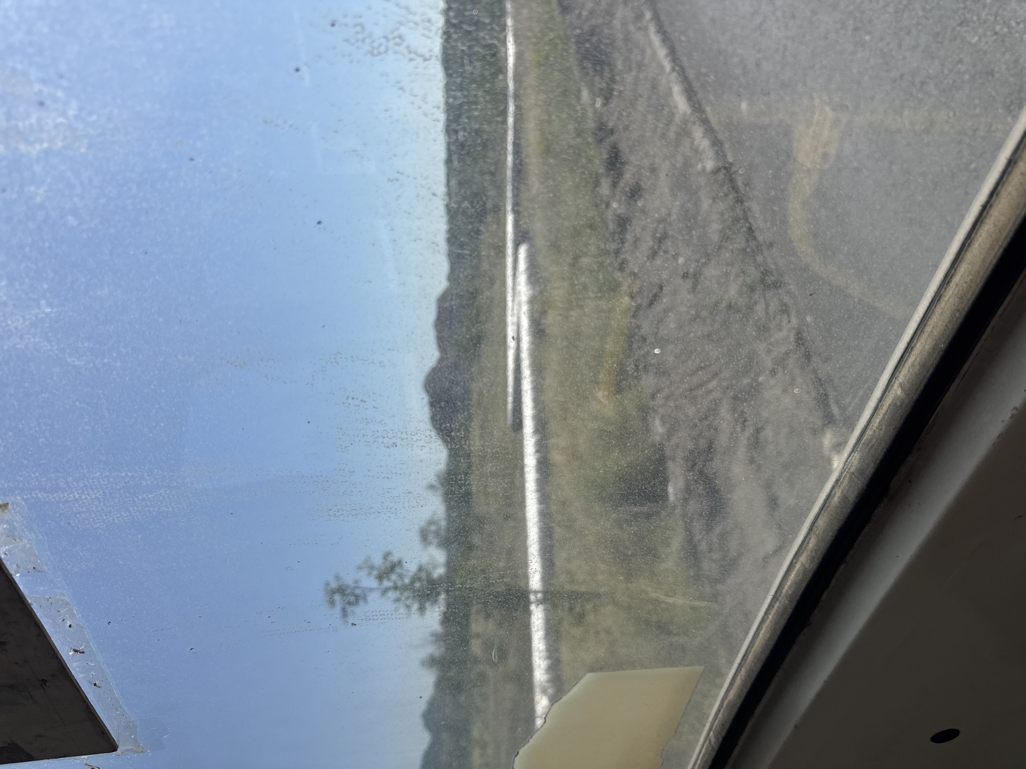 The image shows a view through a dirty window, likely from inside a vehicle. The foreground shows the window frame and a piece of tape on the glass. The background depicts a dry, somewhat barren landscape under a clear blue sky. There are hills in the distance, and a single, sparse tree is visible. A long, reflective pipe or structure runs horizontally across the middle ground, set amidst dry grass and dirt. The overall impression is one of a somewhat desolate, possibly industrial or construction area. The location is not identifiable from the visual information presented.