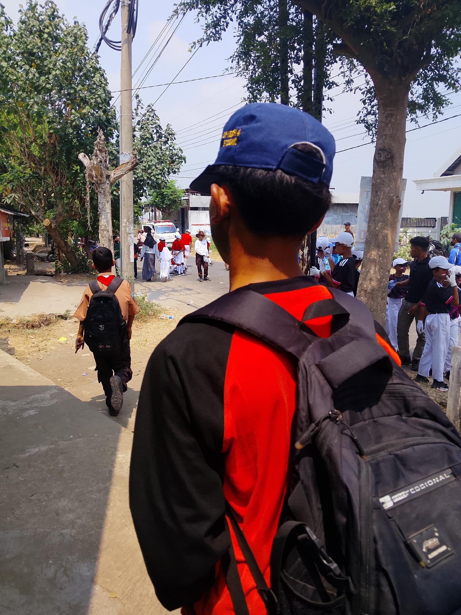In Juwono, Indonesia, under a clear sky, a young boy in a brown scout uniform and black pants with a black backpack walks away from the camera on a dirt and concrete path. He is followed by other children wearing white caps and black and white uniforms, some holding hands. In the background, a white vehicle with red flashing lights and the word "POLISI" is parked. Adults in traditional Islamic attire, including a woman in a black headscarf and a man in a white robe and red sash, are also present. The scene is set against a backdrop of trees, a utility pole with wires, and simple buildings. The foreground is dominated by the back of another person wearing a black and red shirt and a black backpack.