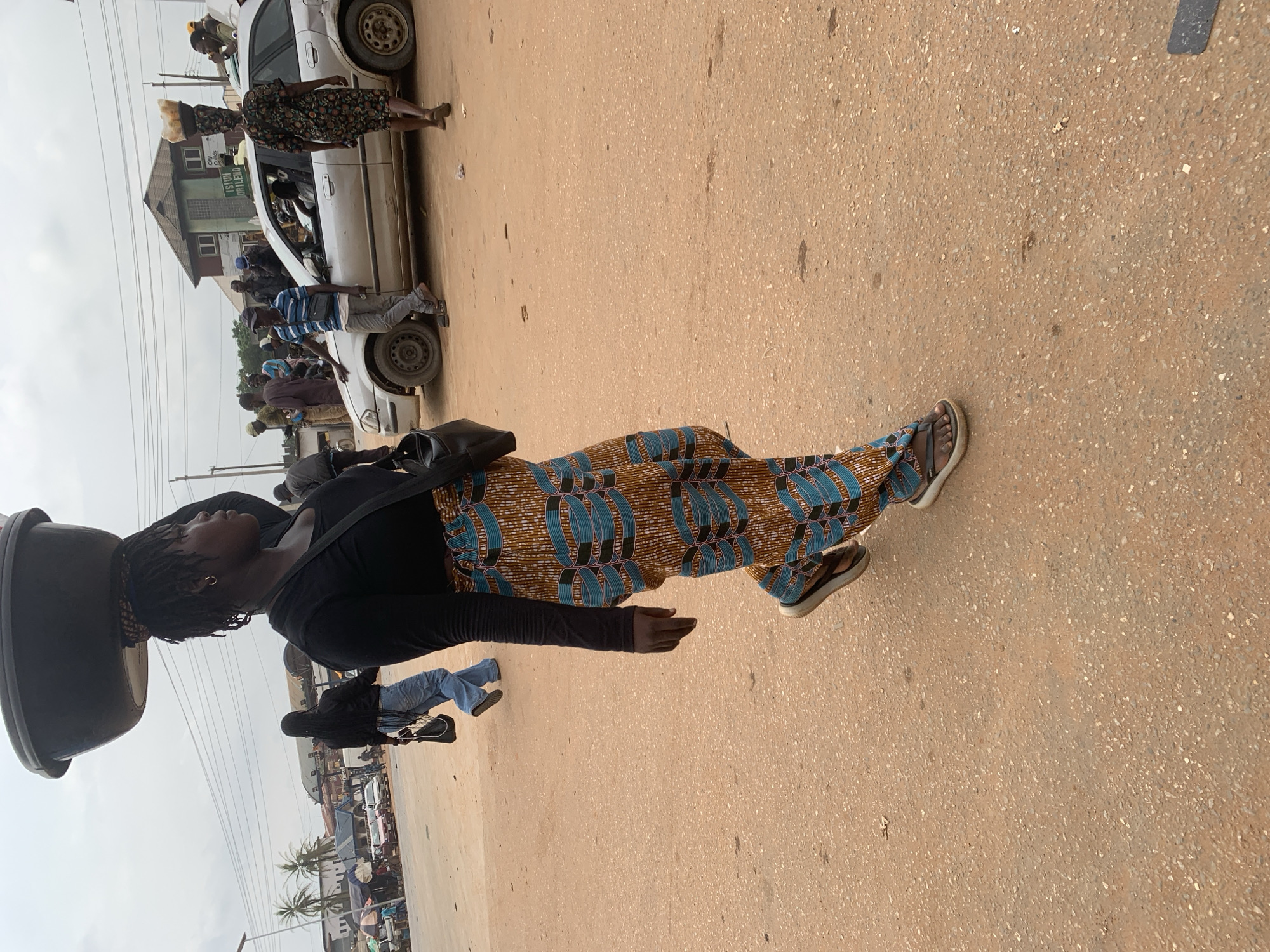 The image depicts a street scene in Shagamu, Nigeria. In the foreground, a woman with dark skin and braided hair walks towards the right, carrying a large dark basin on her head. She is wearing a black long-sleeved top and a brightly patterned skirt with blue and orange designs. She has a dark bag slung over her right shoulder. Her feet are bare except for simple brown sandals. In the background, a silver car is parked, with several people around it. A man in a blue and white striped shirt stands by the driver's side of the car, leaning on it. Another woman, wearing a patterned dress and carrying a basket on her head, walks away from the car. The street is unpaved and appears to be made of reddish-brown dirt mixed with small stones. There are buildings and market stalls lining the street in the distance, and overhead wires are visible against a cloudy sky. The overall impression is of a bustling market environment in a Nigerian city.