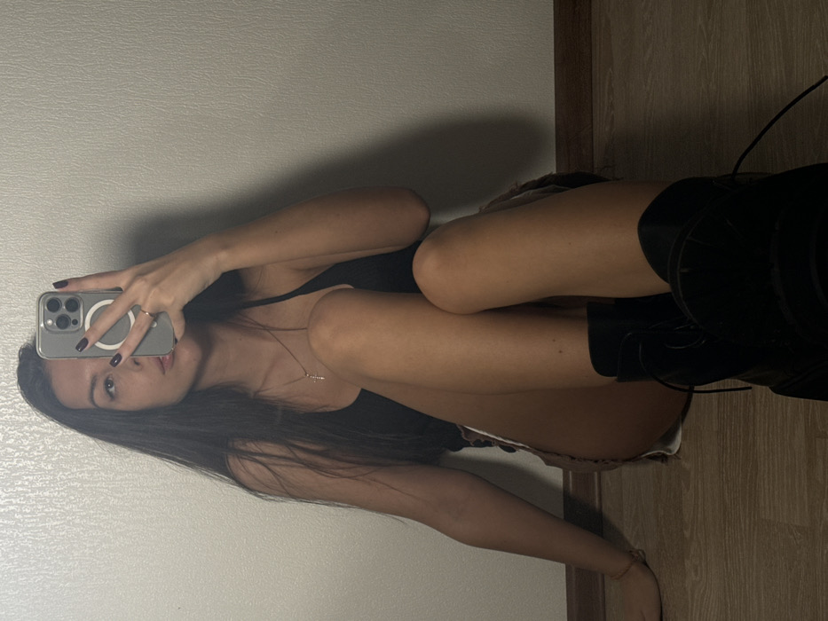 A woman is shown taking a selfie in a mirror. She has long, dark hair and is wearing a black top, shorts, and knee-high black boots with laces. She is holding a smartphone with a clear case and is wearing a ring on her finger. The background consists of a plain white wall and a wooden door frame. The image appears to be taken indoors with natural lighting.