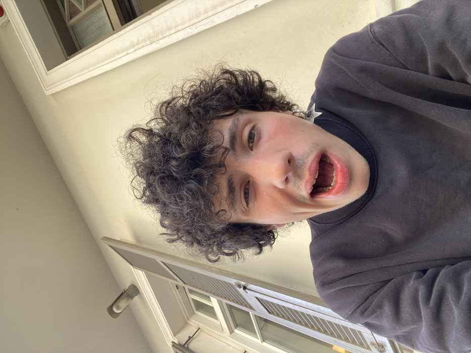 A young man with curly dark hair and a grey sweatshirt stares directly into the camera with his mouth open, possibly shouting. He wears a large silver star earring. The camera angle is low, looking upwards, with a window and building facade visible in the background. The natural light illuminates his face, highlighting his fair skin. The image appears to be a selfie taken outdoors during daylight.