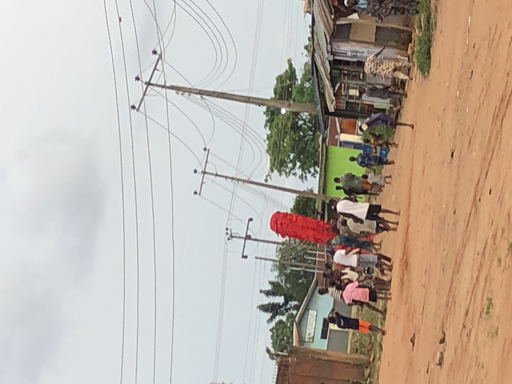 A procession of people moves along a dirt road in Unknown, Unknown. The group includes a large, red, cylindrical object being carried. Overhead power lines stretch across the scene. Buildings flank the road, and trees are visible in the background. The sky is overcast.