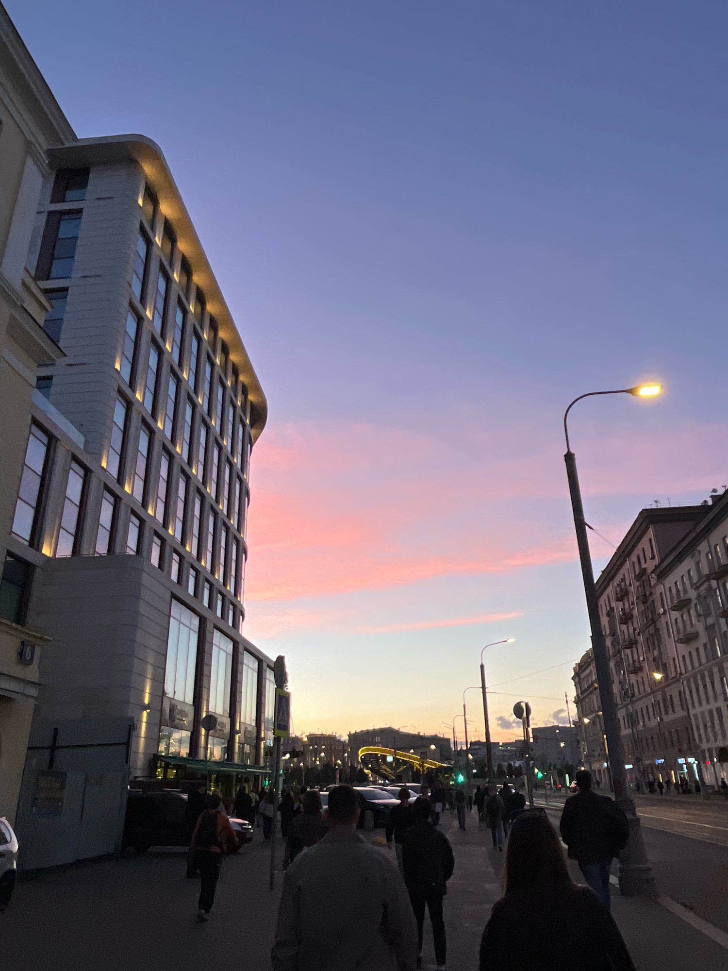 The image depicts a street scene in Moscow, Russia, during twilight. A multi-story modern building with illuminated windows is on the left, adjacent to an older building with more traditional architecture on the right. A street lamp with a lit bulb stands prominently on the sidewalk. Several people are walking along the sidewalk, some in groups, others individually, suggesting pedestrian activity in the urban environment. Vehicles are visible on the street, indicating ongoing traffic. The sky exhibits a gradient of colors, transitioning from a light blue at the top to soft pink and orange hues near the horizon, characteristic of a sunset or sunrise. Traffic lights, also visible, suggest this is a busy intersection or thoroughfare. The overall impression is of a bustling city street transitioning into the evening.