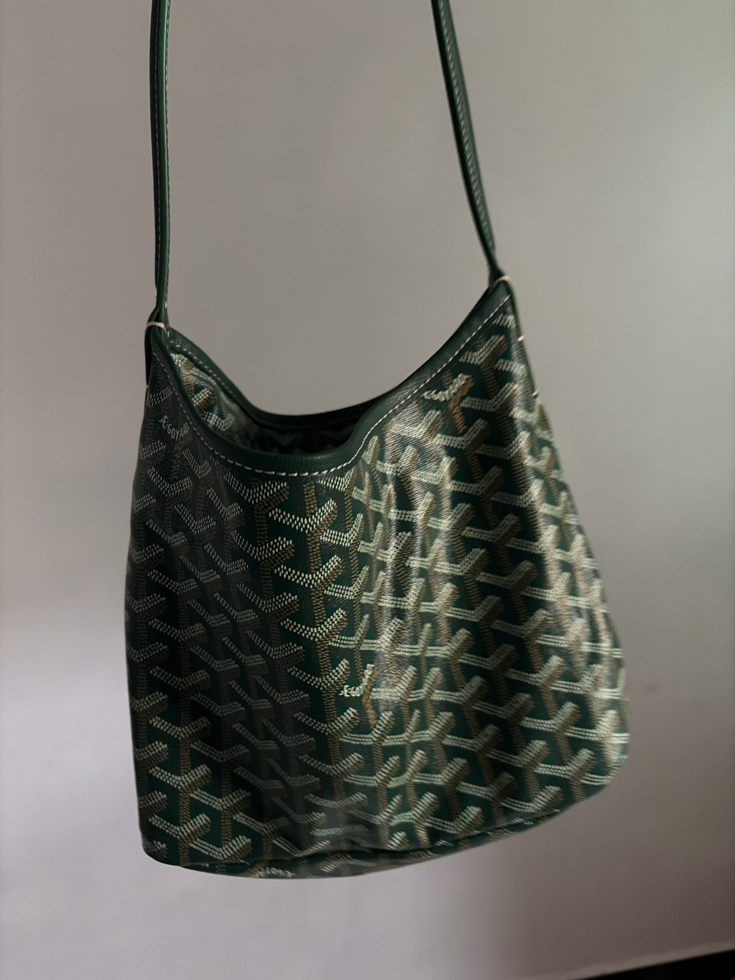 A dark green Goyard shoulder bag hangs suspended against a plain, greyish-white background. The bag features the brand's signature chevron pattern in shades of green and beige. The strap is a dark green, contrasting slightly with the rest of the bag. The lighting casts a subtle shadow to the right of the bag.