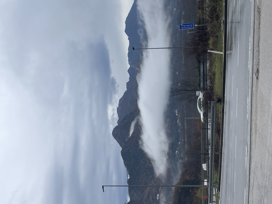 The image captures a mountainous landscape in Sotonići, Montenegro, under an overcast sky. The foreground features a wet, dark asphalt road with white dashed lane markings. A silver metal guardrail runs horizontally across the mid-foreground, separating the road from a strip of grass and sparse, dormant vegetation. A white sedan is partially visible behind the guardrail on the right side, oriented towards the right. Two dark grey streetlights stand upright, one on the left and one on the right, behind the guardrail. A blue rectangular road sign, displaying white arrows indicating a lane merge, is mounted on a pole near the right streetlight. In the midground, a thick, horizontal band of white fog or low cloud extends across the scene, obscuring the lower and mid-sections of the hills and valleys. Small residential structures are faintly discernible beneath the fog on the lower slopes. The background consists of dark, rugged mountains with visible peaks rising above the cloud cover, exhibiting a covering of dark green and brown vegetation. The sky is uniformly grey and heavily clouded. No individuals are present in the scene.