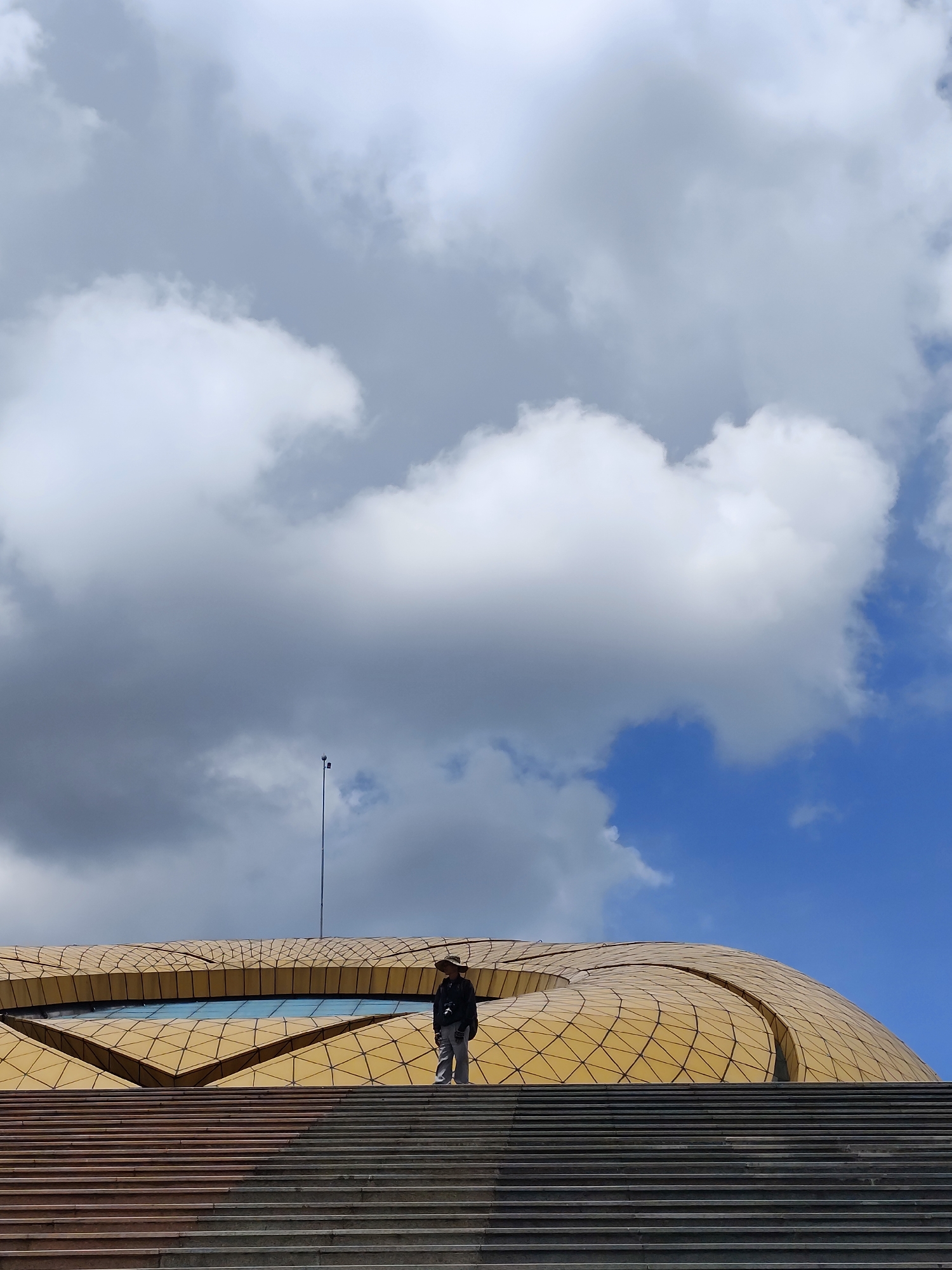 In Da Lat, Vietnam, a person stands on a wide staircase leading up to a modern, golden-colored building with a geometric facade. The sky above is a mix of bright blue and large, dramatic white and gray clouds. The building's roof features an eye-catching, wave-like pattern with a reflective strip that could be glass or polished metal. The person, seen from the waist up, is wearing a hat and dark clothing, and appears to be looking towards the building. The staircase dominates the foreground, with visible lines of concrete or stone steps. A tall, slender pole with two small lights is visible against the cloudy sky. The overall impression is one of architectural grandeur set against a dynamic sky.