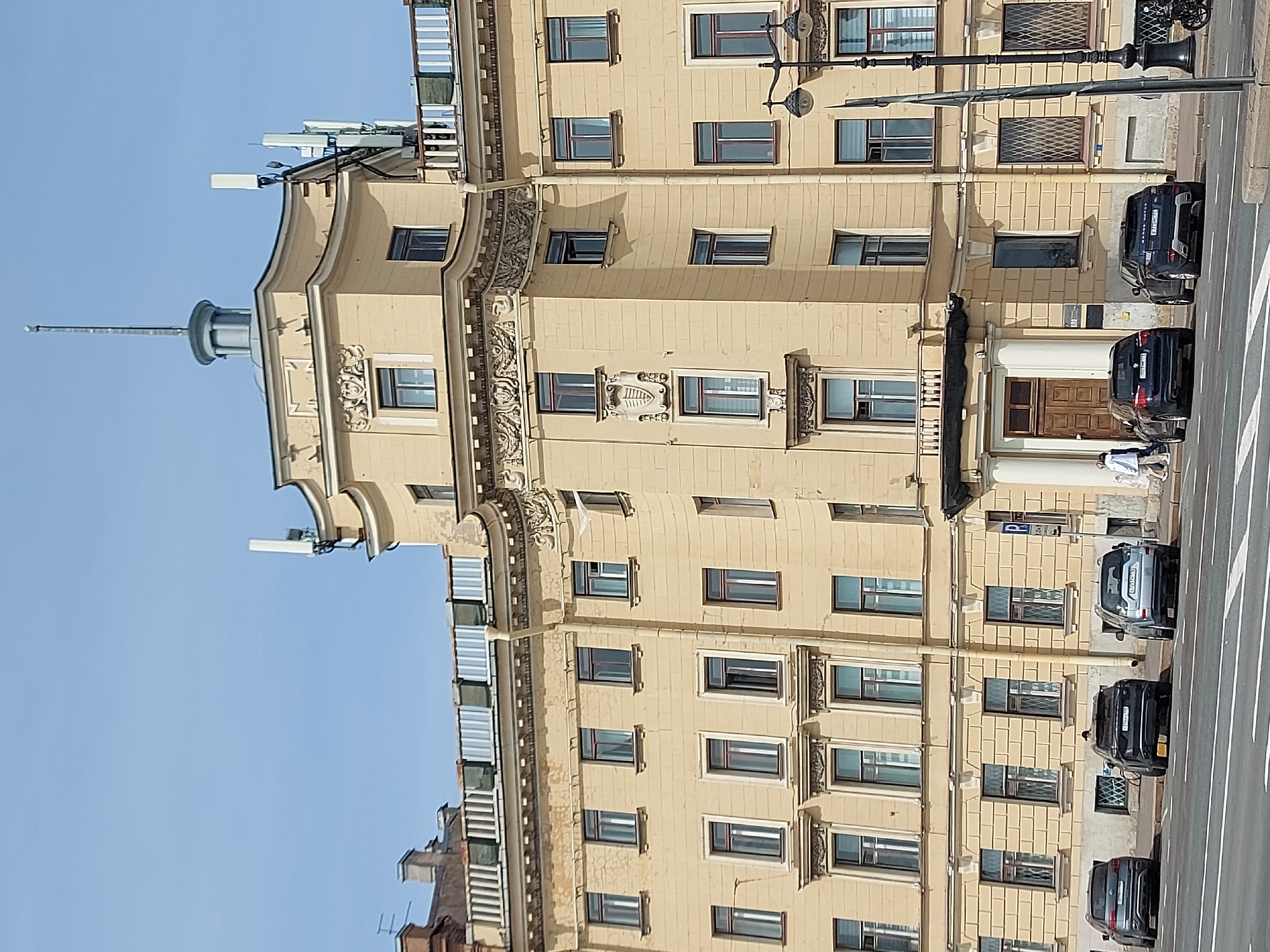 The image captures a stately, light-yellow building in what appears to be Saint Petersburg, Russia. The building is multi-storied, exhibiting numerous windows and ornate architectural details along its facade. A distinct turret-like structure is visible atop one corner. A street runs alongside the building, featuring several parked vehicles. Pedestrians are present on the sidewalk near the building's entrance. Street lamps are also evident along the road. The sky above is clear and blue, indicating a sunny day.