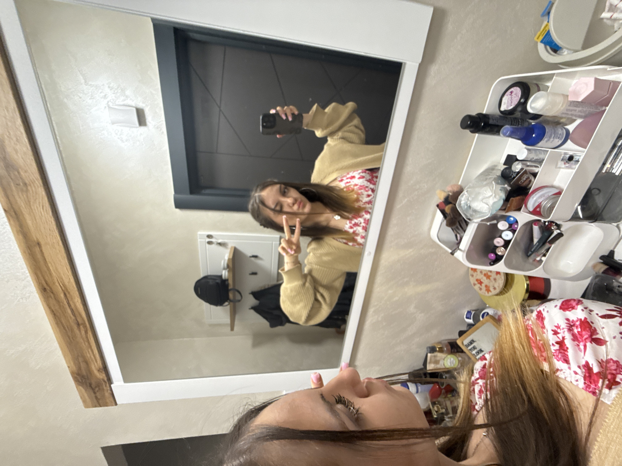 The image shows a young woman taking a selfie in a mirror. She is wearing a floral dress and a beige cardigan. Her hair is brown. She is holding up two fingers in a peace sign. The background of the mirror shows a door, a coat rack, and a backpack. To the right of the mirror is a makeup organizer filled with various cosmetics. The location appears to be indoors, possibly in Sofia, Bulgaria.