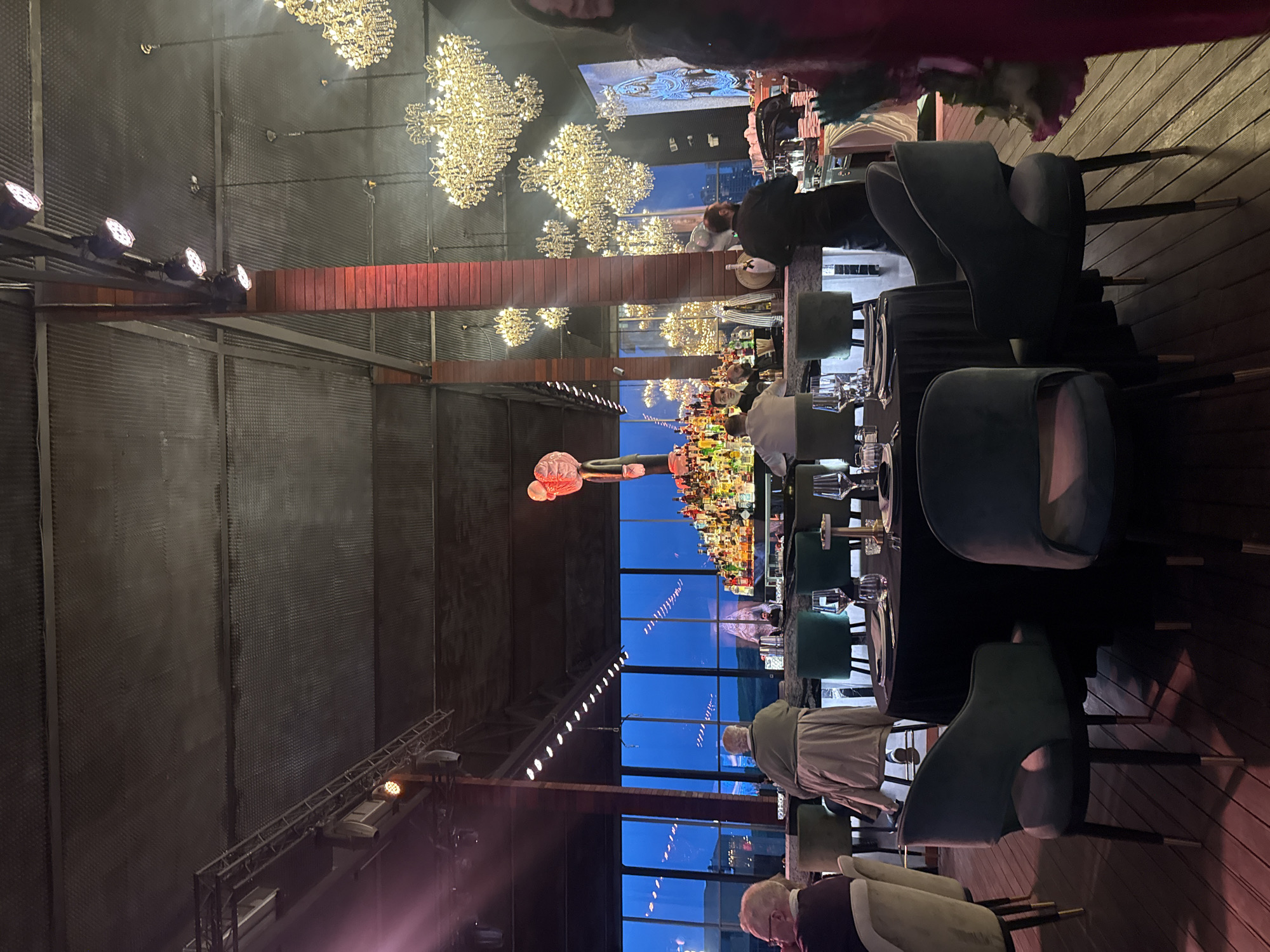 The image shows the interior of a high-end restaurant or event space in Yerevan, Armenia, captured during the evening or nighttime.
Environment: The venue features a high ceiling with exposed industrial-style beams and trusses. Rows of decorative chandeliers provide ambient lighting. Large windows provide a view of the cityscape at dusk or night, with dark blue skies visible.
Objects: The scene is set for dining, with tables covered in black tablecloths and chairs with a distinctive teal-green upholstery. Each place setting is complete with silverware, glasses, and plates, indicating a formal dinner or event setup. In the middle of the room, a long table is adorned with what appears to be an elaborate floral arrangement or a decorated centerpiece, and in the back of the room, there is an elaborately stocked bar.
People: Several people are present. Some are seated at the tables. There are people standing behind the bar, presumably employees or guests. They are dressed in a variety of attire.
Actions/Interactions: The people appear to be either preparing for an event, conversing, or waiting to be seated.
Overall: The scene depicts a sophisticated, modern restaurant in Yerevan ready for an evening event.