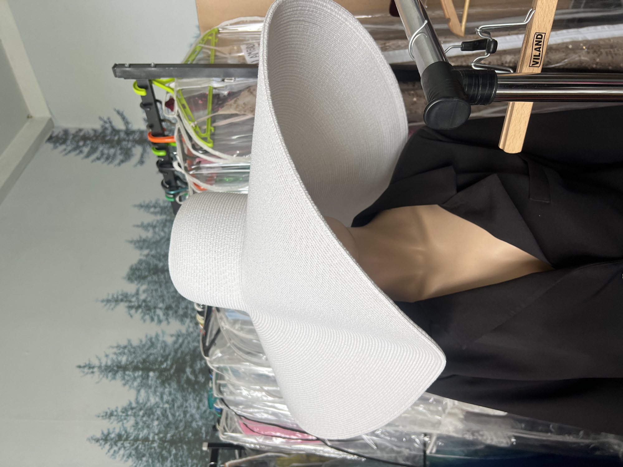 The image shows a white, wide-brimmed sun hat on a mannequin, in what appears to be a clothing store. The mannequin is wearing a black blazer. In the background, there are racks of clothing, some of which are covered in plastic garment bags. A mural of pine trees is visible on the wall behind the racks. A wooden hanger with the word "VILAND" is also visible. The lighting suggests an indoor setting.