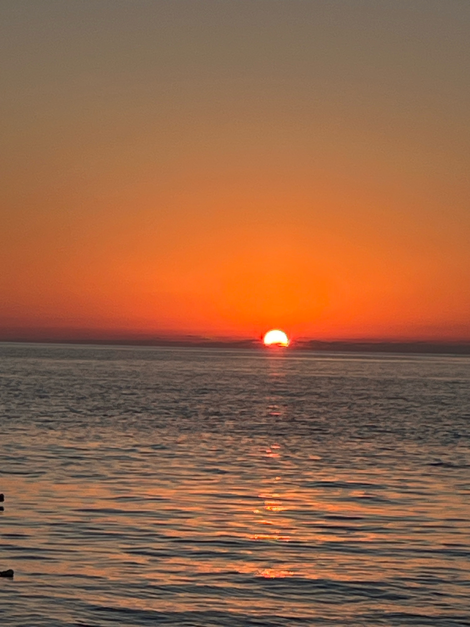 The image captures a serene sunset over the Black Sea in Kobuleti, Georgia. The sky is a gradient of warm orange and yellow hues, transitioning to a muted gray at the top of the frame. The sun, a vibrant orange disc, is partially submerged at the horizon, casting a shimmering reflection on the calm water. The water itself appears dark with gentle ripples, reflecting the warm colors of the sky. The overall scene is one of peaceful natural beauty.
