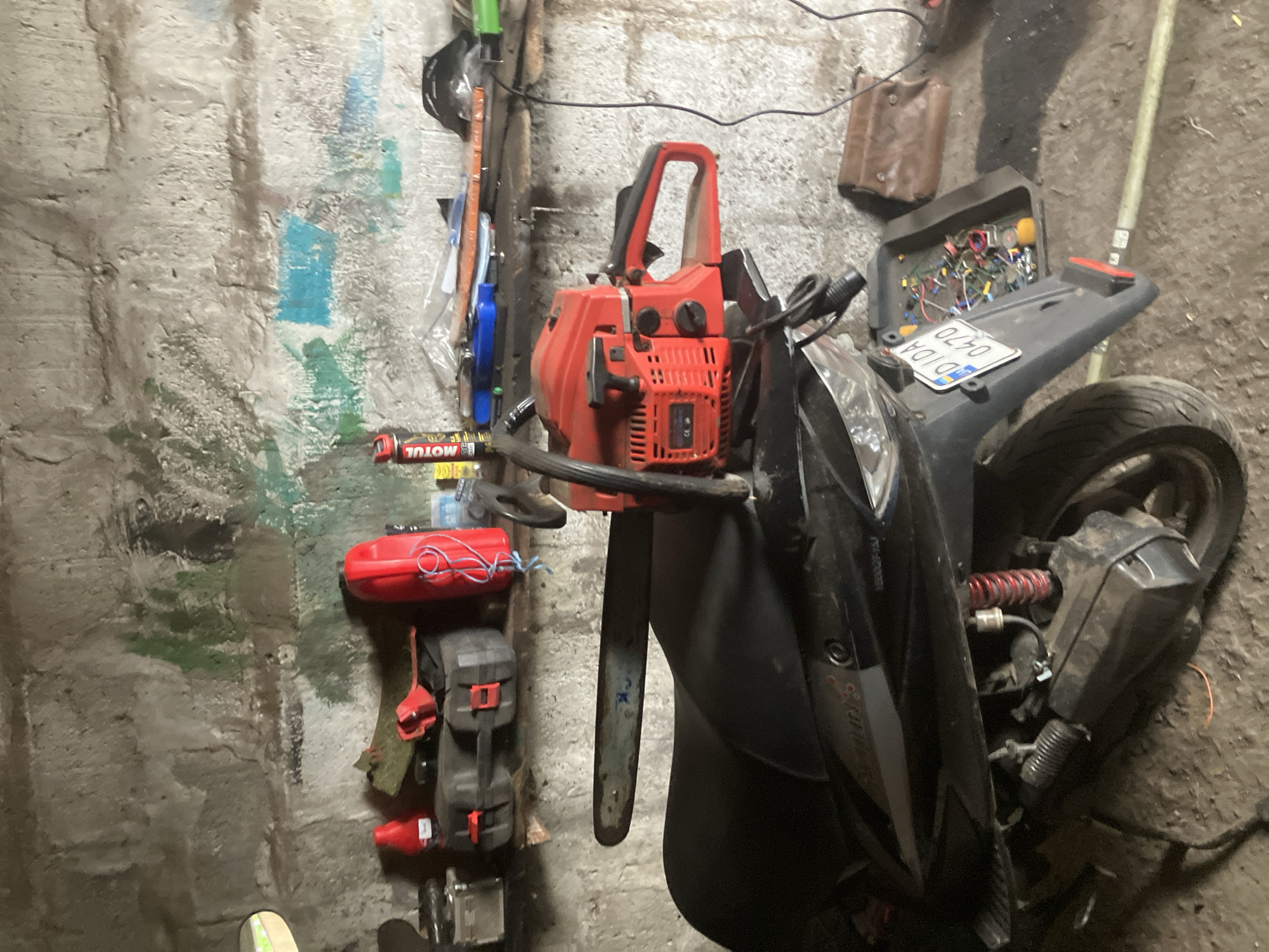 In a dimly lit, possibly residential garage or workshop in Vovkivka, Ukraine, a red and black chainsaw is prominently placed across the seat of a black scooter. The scooter has a license plate with the characters "DDIDA 0470". To the left of the scooter, on a shelf against a rough, textured wall, are various items including a red fuel can, a black toolbox with red latches, and a can of "MOTUL" oil. Further to the right on the shelf, there are other assorted objects and what appears to be a small brown leather pouch on the floor. The overall environment suggests a storage or work area with a collection of tools and vehicle-related items. There are no people visible in the image.