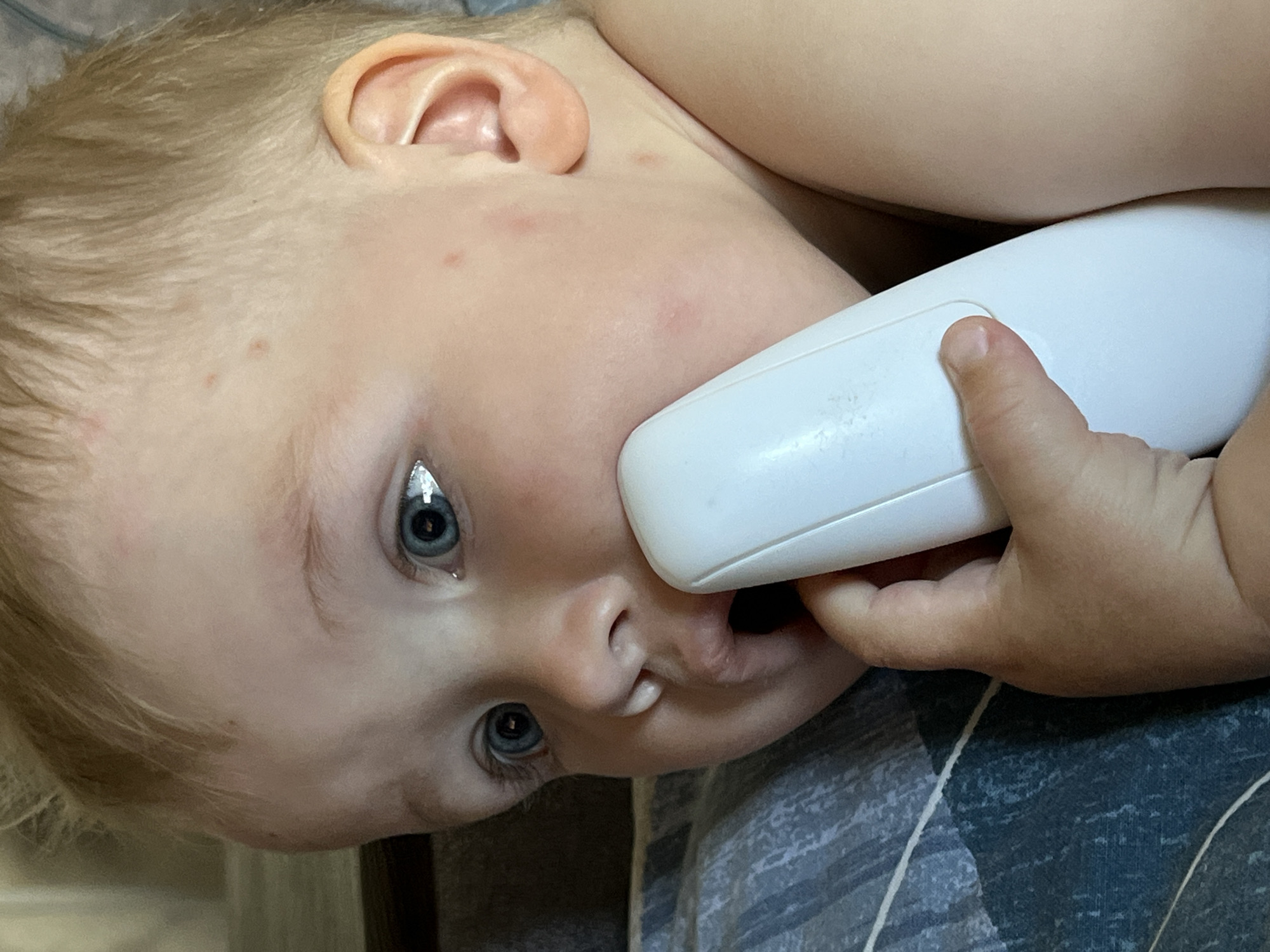 A blond-haired toddler with blue eyes is lying down, holding a white object to their face. The object appears to be an electronic device. The child's left ear is visible and there are a few small red spots on their face. The child is lying on a blue and grey patterned surface. The location and city cannot be determined from the image.