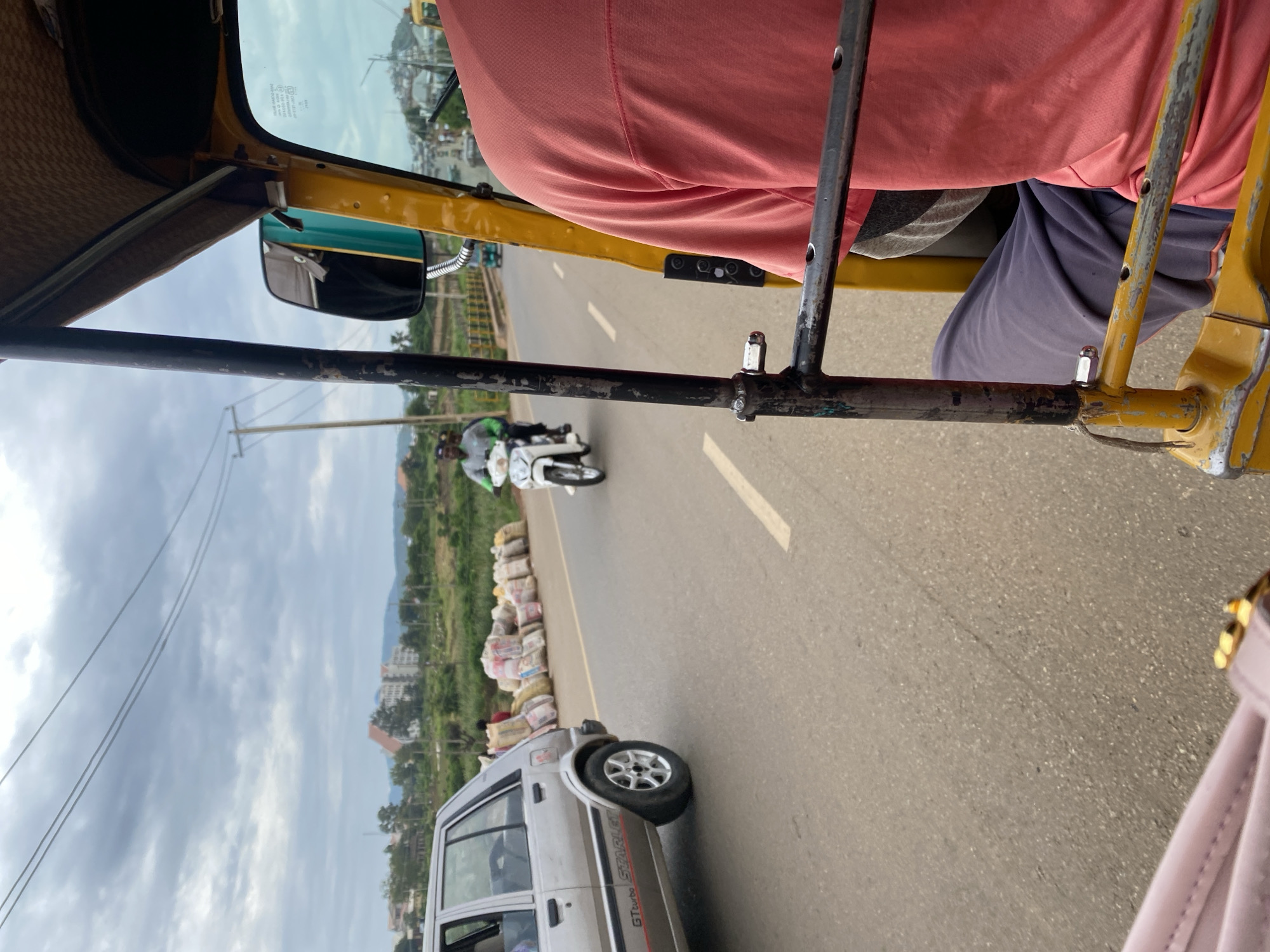 The image shows a view from the passenger seat of a yellow auto-rickshaw driving on a paved road. The auto-rickshaw has a straw-like sunshade and a black metal frame. A person wearing a pink shirt and blue pants is visible in the passenger seat. In front of the auto-rickshaw, a motorcycle carrying two people is seen on the road. The motorcycle rider is wearing a helmet and a green shirt, and the passenger is wearing a white shirt.
On the side of the road, there are many sacks stacked up, possibly containing goods or produce. Further down the road, a silver car is parked. Beyond the road, there are buildings, trees, and a cloudy sky. The scene appears to be in a town or city in Nigeria, as suggested by the location context "Rafin Par, Nigeria". The sky is overcast, indicating cloudy weather. The time of day is likely daytime, given the lighting conditions. The overall mood is that of everyday travel and commerce. There is no visible text in the image.
