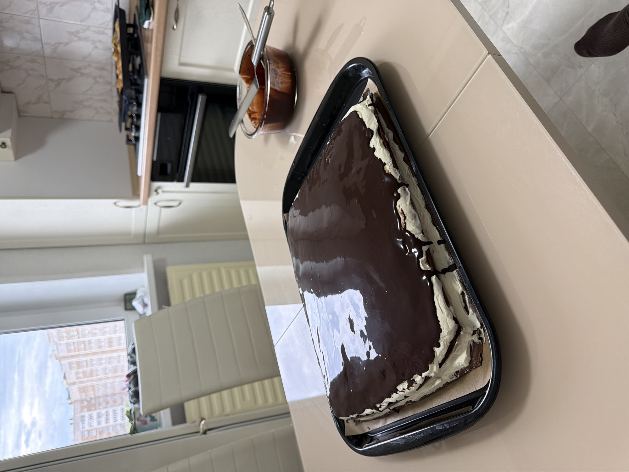 This image captures a domestic kitchen scene, likely in Izhevsk, Russia, focused on a freshly prepared chocolate cake.
The primary subject is a large, rectangular cake, generously coated with a glossy, dark chocolate glaze, resting on a black baking tray lined with parchment paper. White cream or frosting is visible along the sides of the cake, peeking out from beneath the dark glaze. Adjacent to the cake, further back on the light beige countertop, a small, clear pot holds residual dark chocolate glaze, with a metal spatula or spoon resting within it, suggesting recent use.
In the background to the left, a section of a modern kitchen is visible, featuring white cabinets, a built-in microwave oven, and part of a stovetop. A window dominates the left side of the frame, offering a view of a multi-story urban building with numerous windows and a light-colored facade, suggesting a residential area. The sky outside appears somewhat overcast, indicating daytime. Below the window, a white radiator and partially drawn blinds are visible. In the upper right corner, a small portion of a tiled floor and a person's foot clad in a dark sock can be seen.
The activity taking place is the finishing of a baked good, specifically the glazing of a cake, implying home baking. The overall setting is a clean and functional kitchen. There is no visible text in the image.