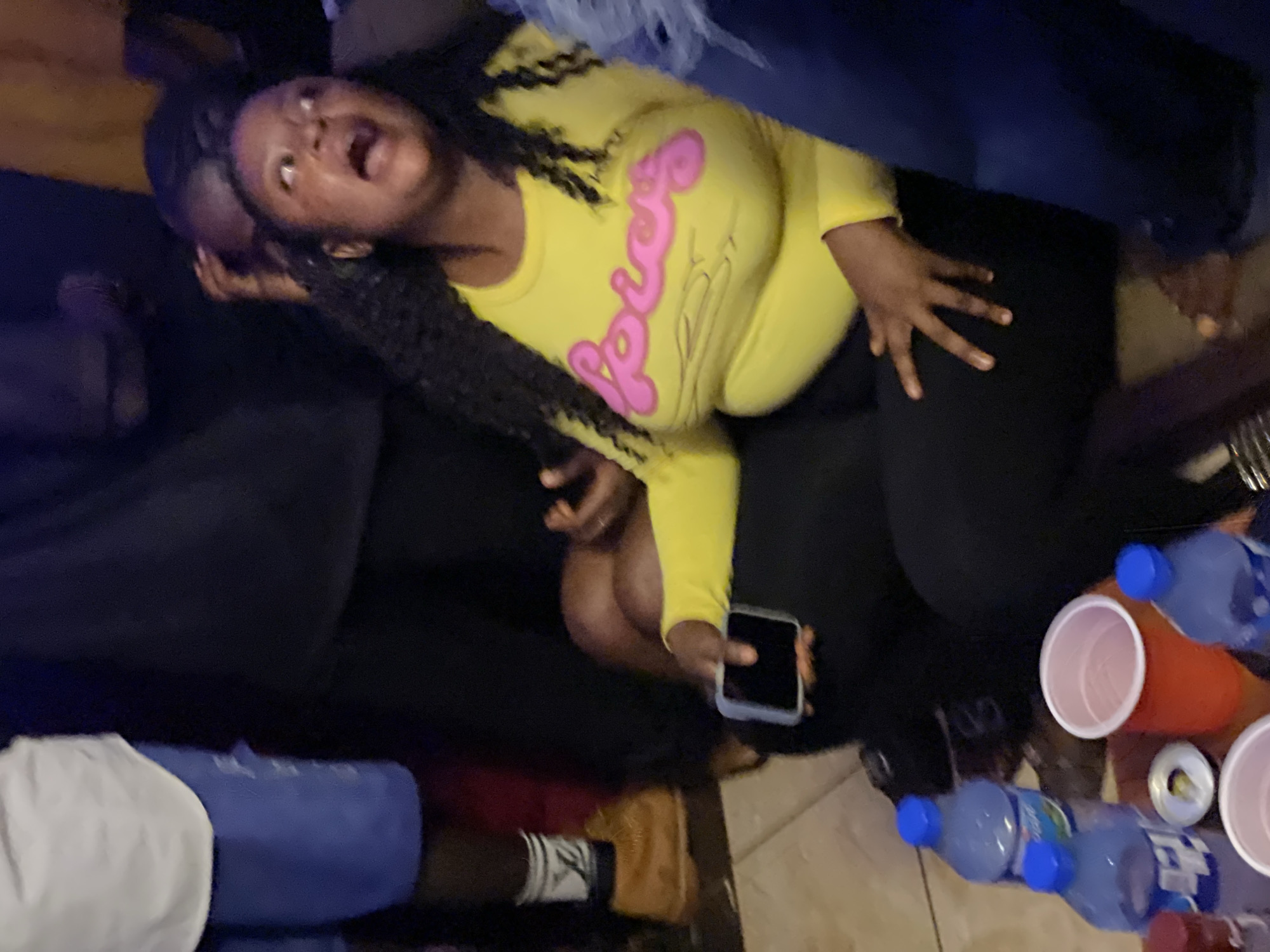 In a dimly lit indoor environment, a young Black woman is the central focus. She is seated on the floor, wearing a bright yellow long-sleeved shirt with "Spicy" written in pink bubble letters across the chest, and black leggings. Her mouth is open, and her eyes are wide, suggesting a moment of surprise or excitement. To her left, her hand rests on a black smartphone. In the foreground, several blue water bottles with white labels and red plastic cups are arranged on a surface. The floor appears to be tiled. The scene suggests a casual gathering or party, potentially in a home or a relaxed social space within Agwala, Nigeria.