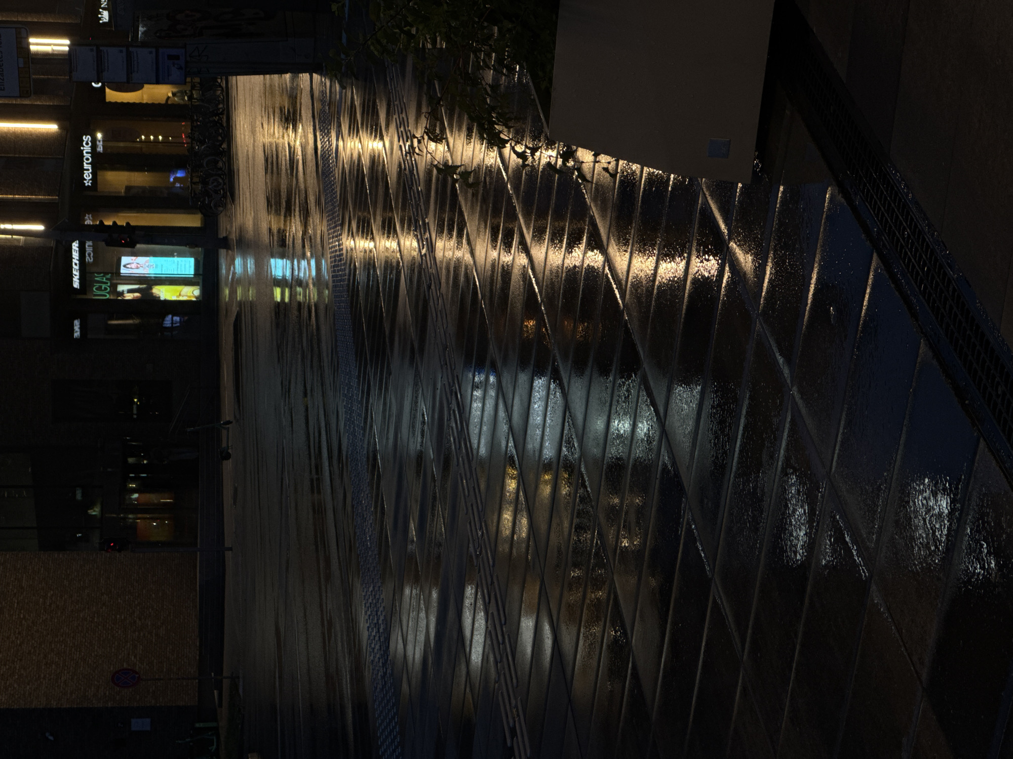 The image depicts a wet, tiled sidewalk at night in Riga, Latvia. The tiles are dark and reflective, with light from storefronts creating shimmering patterns on the wet surface. In the background, storefronts with illuminated signs are visible, including "SKECHERS" and "euronics". A traffic light is also visible. There are no people clearly depicted in the image. Bicycles are parked in front of the storefronts, and an electric scooter is on the sidewalk. A red circular traffic sign with a diagonal line through it is on the left side of the image. A planter with greenery is present on the right side of the image.