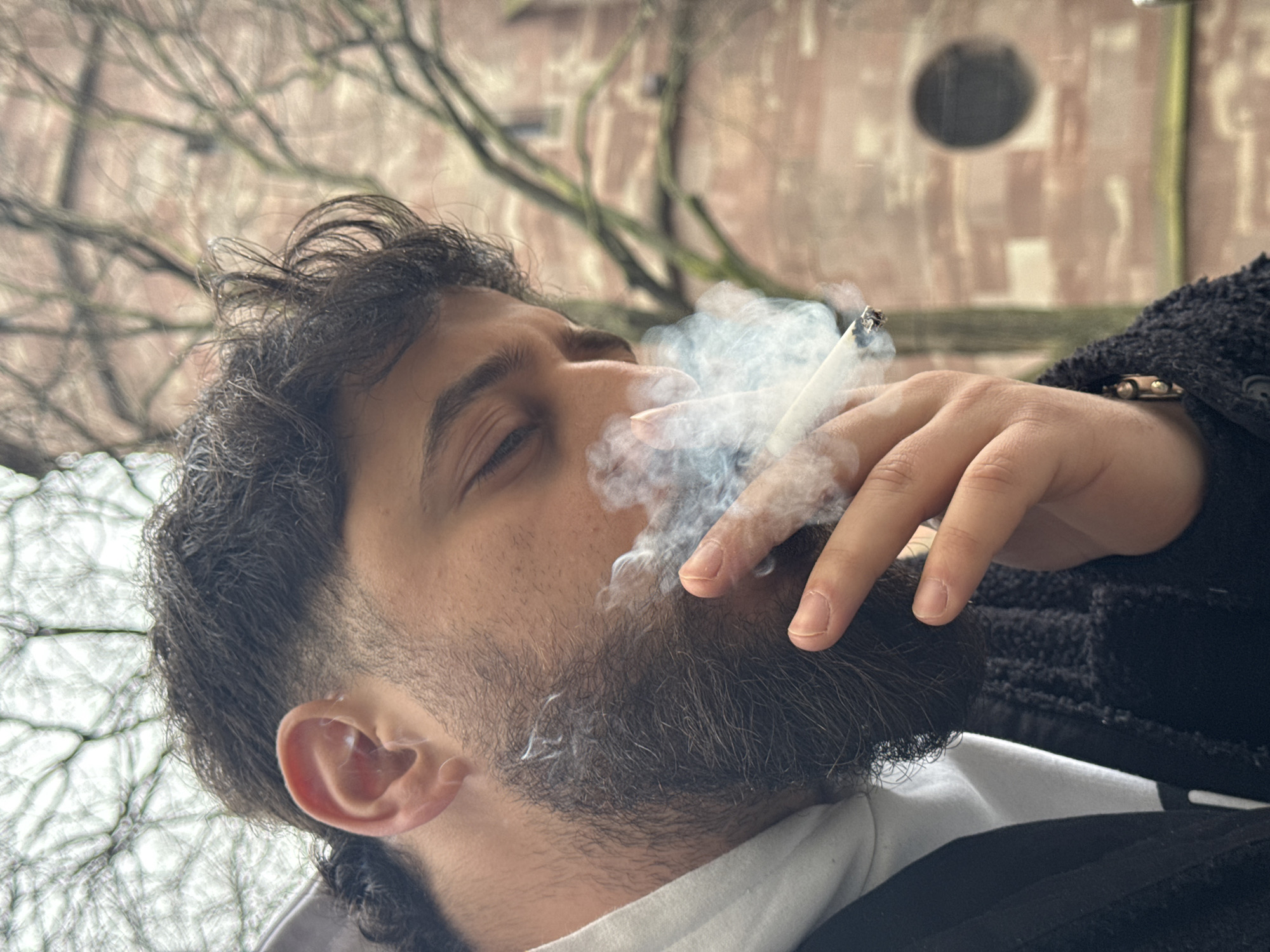 A male individual is depicted in a close-up outdoor scene, exhaling a plume of smoke from a lit cigarette held between his right index finger and thumb. He possesses dark, curly hair with a faded cut on the sides, and a full, dark beard. His eyes are directed downwards or closed. He wears a white hooded garment beneath a dark, textured jacket, with a dark button visible on the jacket's front. A bracelet is visible on his right wrist. The background is out of focus, featuring bare tree branches against an overcast sky. Behind the foliage, a building constructed of reddish-brown stone or brick is visible, characterized by an archway or window and a prominent dark circular feature on its facade. The architectural style and material are characteristic of historic structures in Strasbourg, France, specifically consistent with buildings found in the vicinity of the Cathédrale Notre-Dame de Strasbourg.