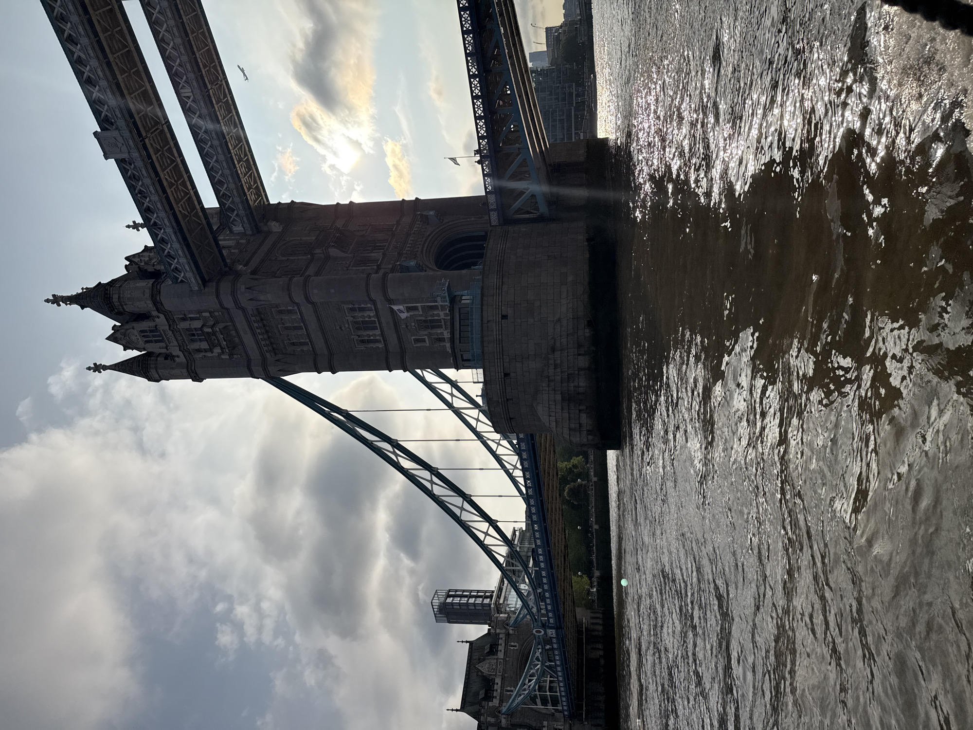 This image features the Tower Bridge in London, United Kingdom. The bridge's architectural details, including its towers, arches, and suspension elements, are visible. The River Thames is also depicted with sunlight reflecting off the water. A cloudy sky forms the backdrop of the scene.