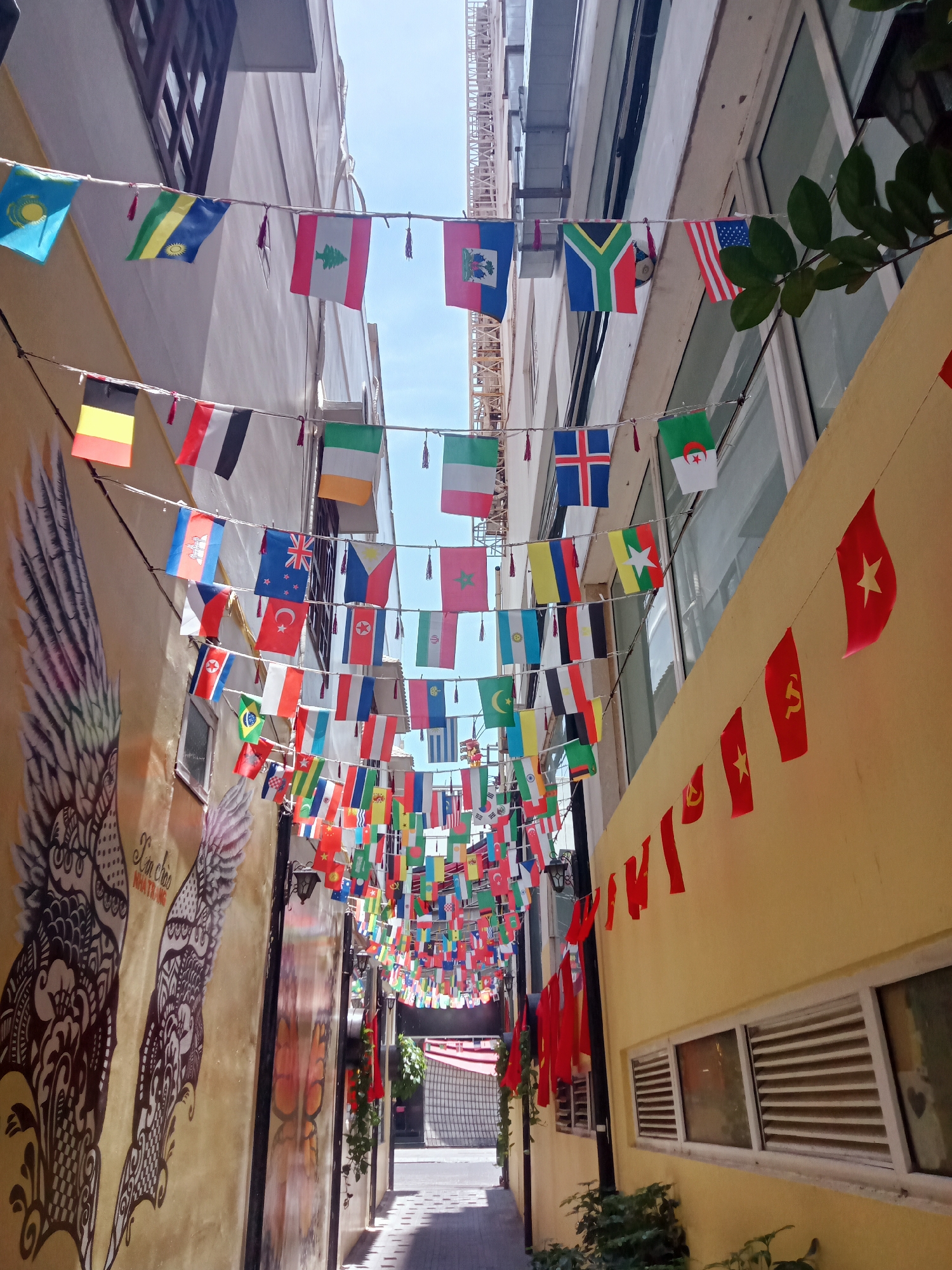 An alleyway in Nha Trang, Vietnam, is decorated with strings of international flags and Vietnamese communist flags. The alley is narrow, with buildings on either side, one with a large mural of a winged figure, the other painted yellow with windows. The strings of flags are hung overhead, creating a colorful canopy. At the end of the alley is a red awning.