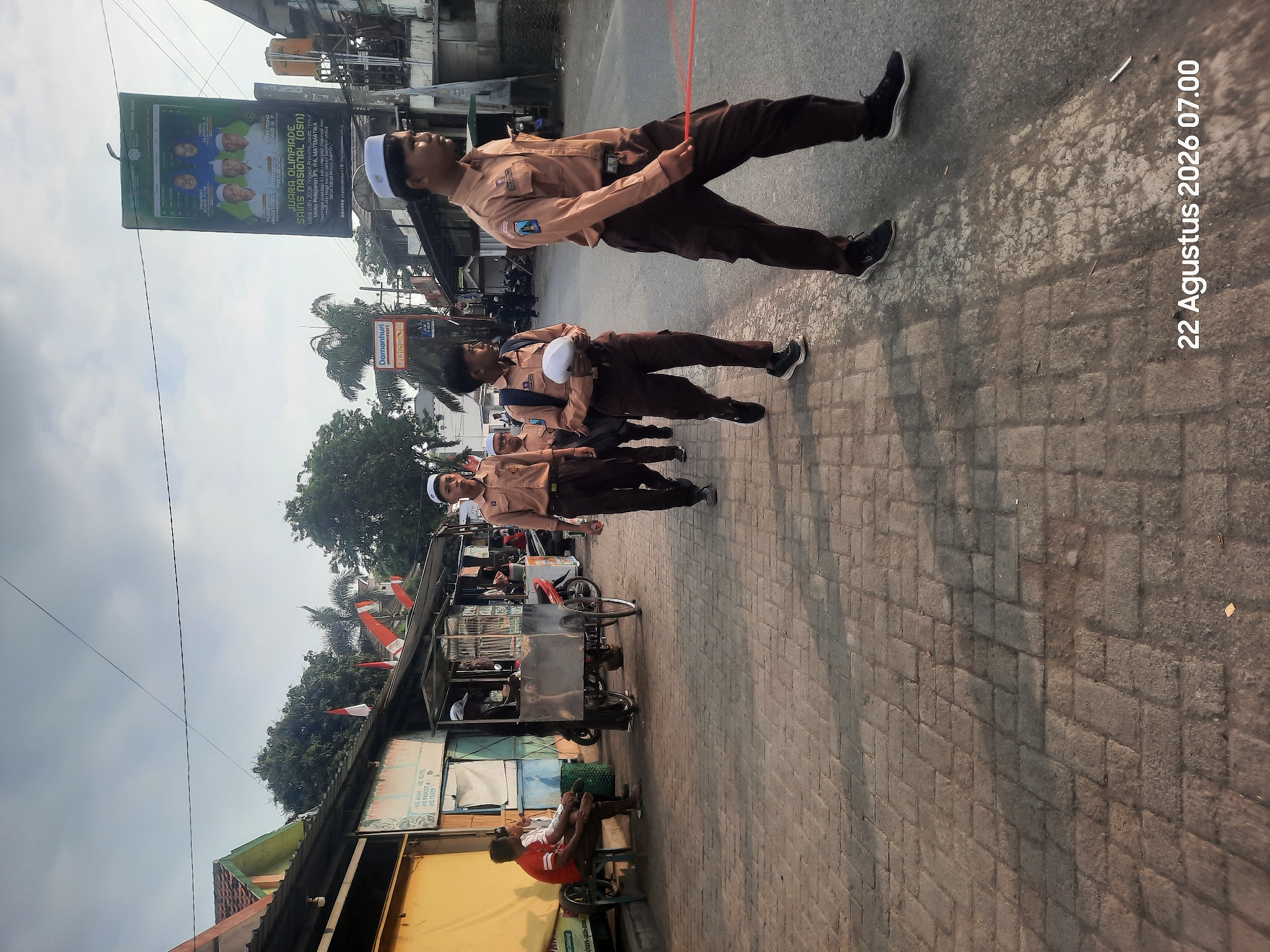 The image depicts a street scene in Pajarakan Kulon, Indonesia, under a cloudy sky. A group of students in light brown uniforms and white skullcaps are walking along a paved sidewalk. One student in the foreground is carrying a red ribbon. To the left of the sidewalk, there are several small shops with awnings and an outdoor seating area where two men are sitting. A food cart is parked on the sidewalk. Further down the street, a banner advertising an academic competition is displayed. The overall atmosphere suggests a normal day in a residential or commercial area of the city.