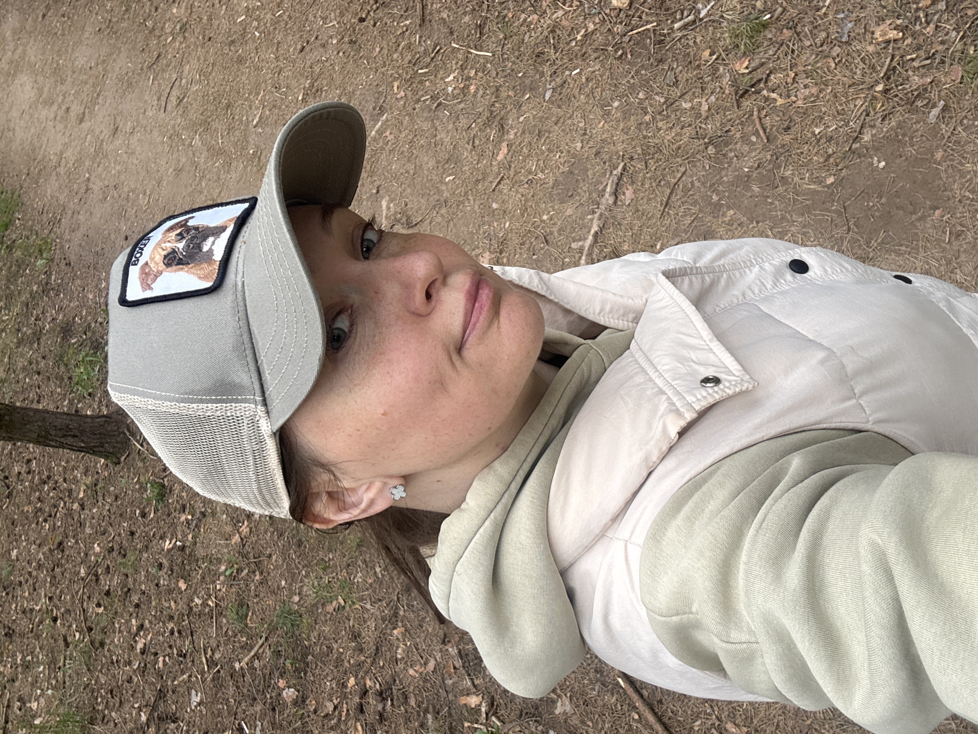 A selfie-style photograph shows a fair-skinned woman with light eyes and light brown hair wearing a light gray baseball cap with a patch of a boxer dog on the front. She is also wearing a light green hooded sweatshirt and a light beige vest. The background appears to be an outdoor area with a dirt path and fallen leaves and twigs on the ground. The image is oriented in a way that suggests the photograph was taken while looking down at the subject. Without additional geographic data, it's impossible to confirm or deny if the image was taken in Chaikovsky, Russia.