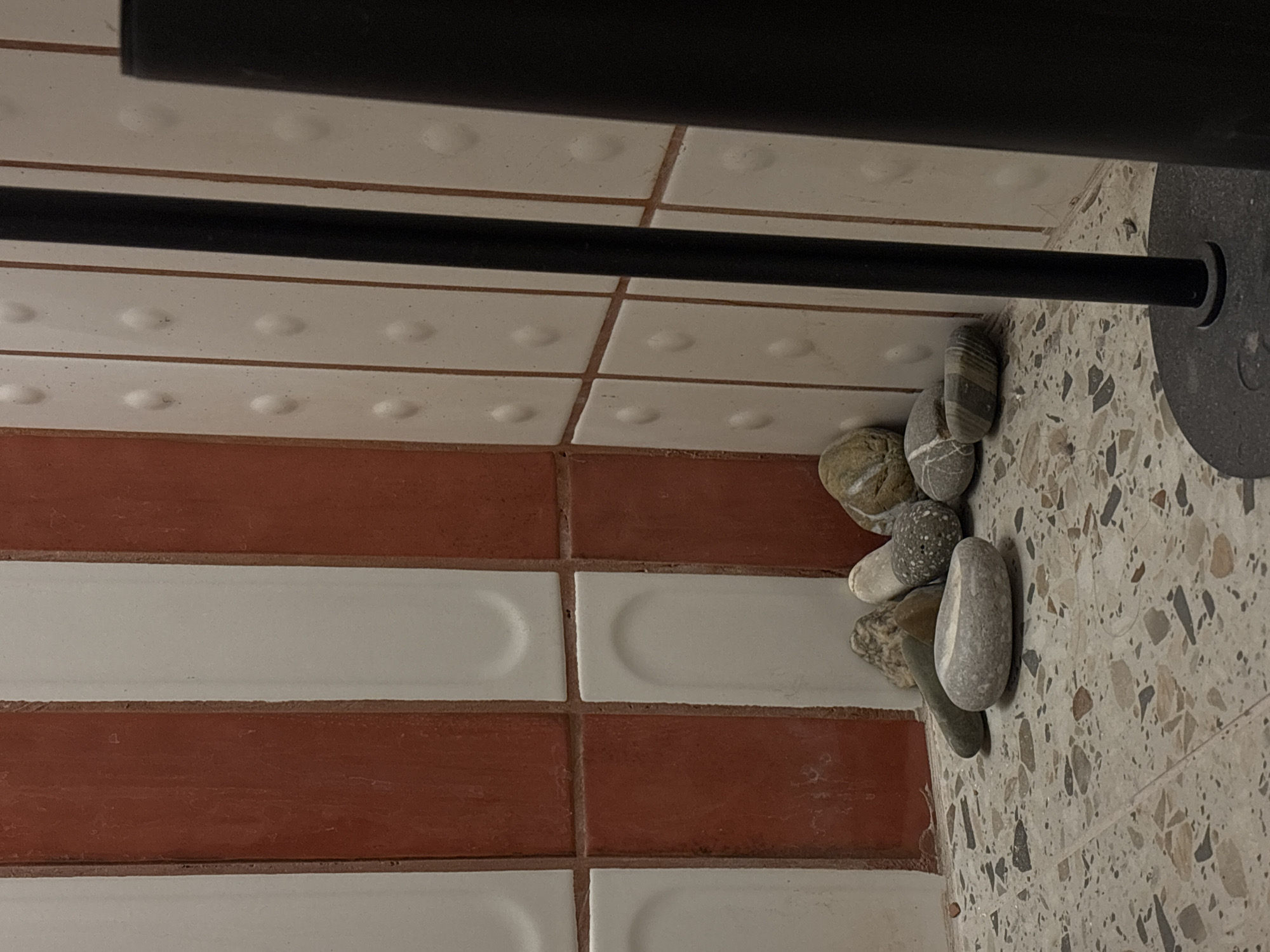 The image shows a section of tiled flooring with a black metal object running across the top and left sides of the frame. The floor is composed of alternating rows of red and white rectangular tiles. Additionally, there are square white tiles with raised circular bumps in the upper part of the image. A collection of grey and brown pebbles is nestled against the edge where the tiles meet a patterned floor. The patterned floor appears to be light grey with dark grey and brown speckles. The lower right corner is dominated by the patterned floor, while the tiled area is more prominent on the left side. The presence of the different tile types and pebbles alongside a metal object suggests an interior space, such as a building with varied flooring designs. No people, actions, or interactions are observed in the scene. Given the provided location of Krasnoyarsk, Russia, the scene could be a room or part of a building in that city.
