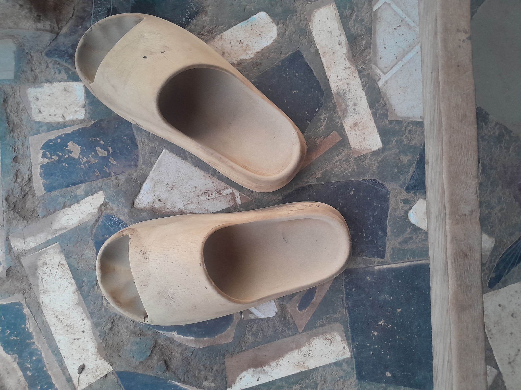 This image depicts two beige slide sandals resting on a patterned tiled ground in Ifo, Nigeria. The sandal on the left is oriented vertically, toe pointing towards the top of the frame. The sandal on the right is diagonally oriented, with its toe pointing towards the upper right. Both sandals show signs of wear, including dirt accumulation and minor surface abrasions. The ground consists of an arrangement of irregularly shaped tiles in various shades of dark blue, grey, and off-white, also exhibiting dirt and wear. A weathered, light brown wooden plank or beam extends horizontally across the lower portion of the image. No individuals, actions, or interactions are present in the scene.